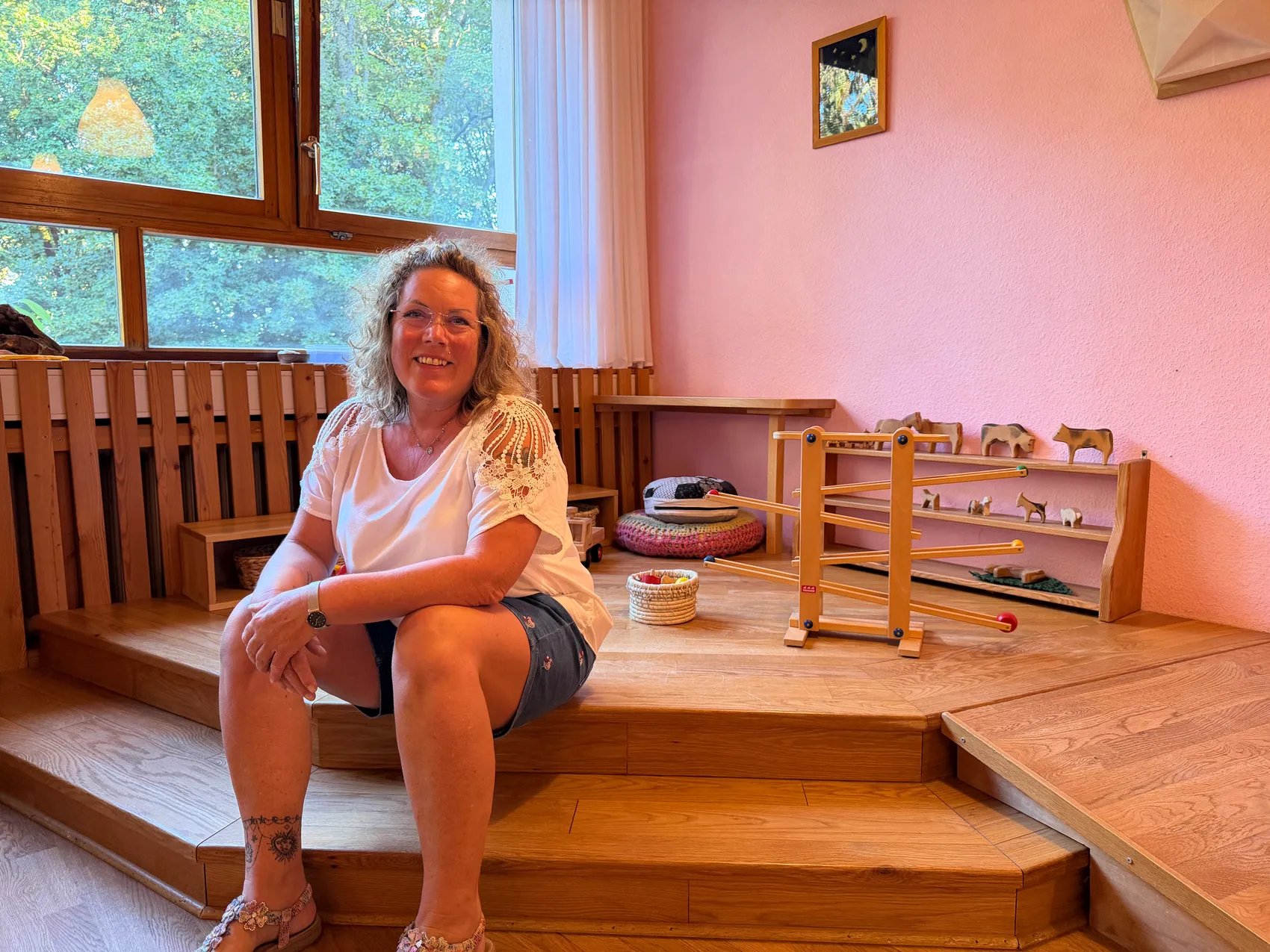 Ananda Schüller sitzt lächelnd auf einer Holztreppe in einem hellen Raum mit rosa Wänden und Holzspielzeug.