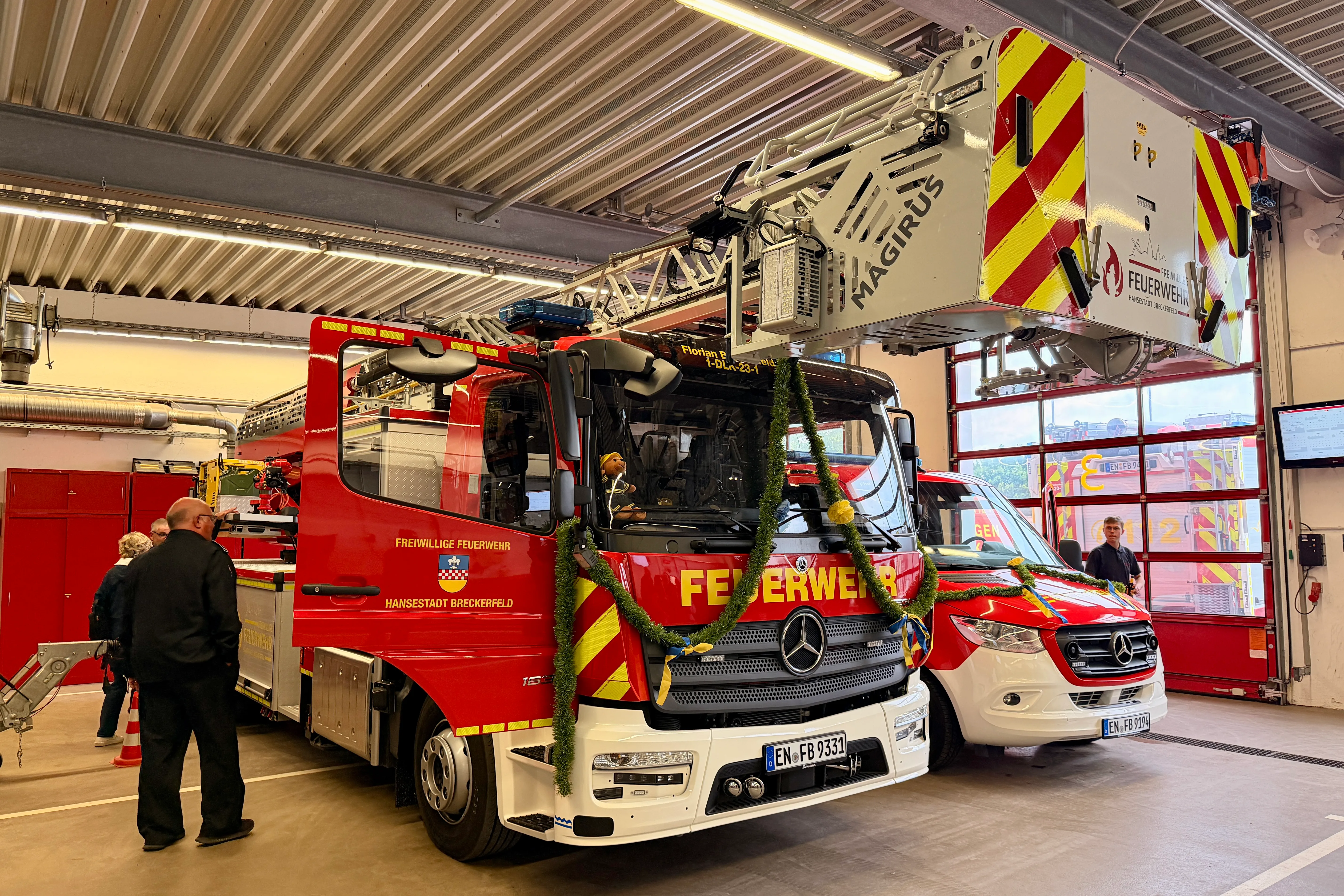 Ein roter Feuerwehrauto mit Girlanden steht in einer Feuerwache, umgeben von Feuerwehrleuten und einem weiteren weißen Fahrzeug. Im Hintergrund ist ein Bildschirm mit dem Schriftzug „Feuerwehr“ zu sehen.