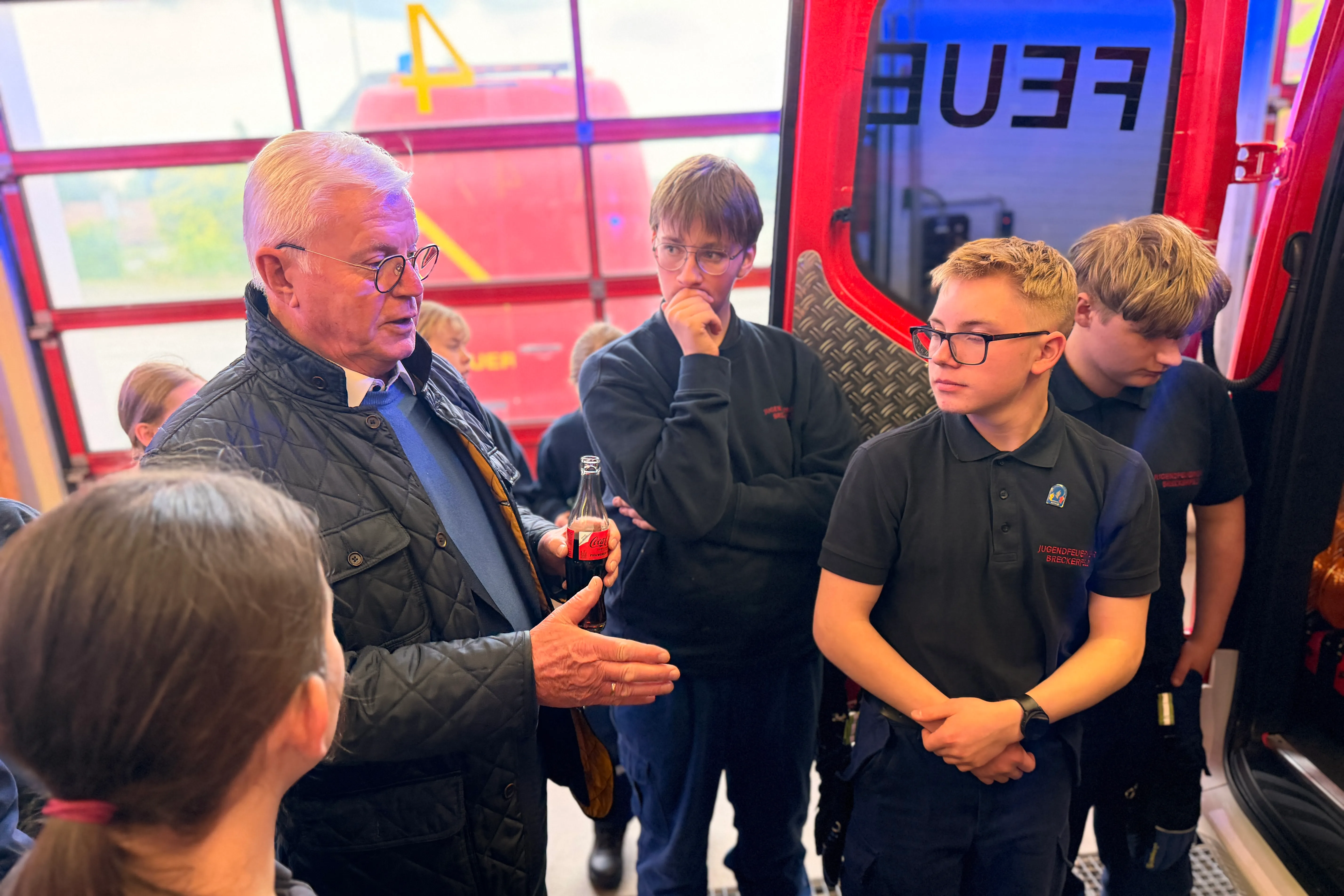 Ein älterer Mann mit Brille erklärt Jugendlichen die Technik eines Feuerwehrautos im Hintergrund.