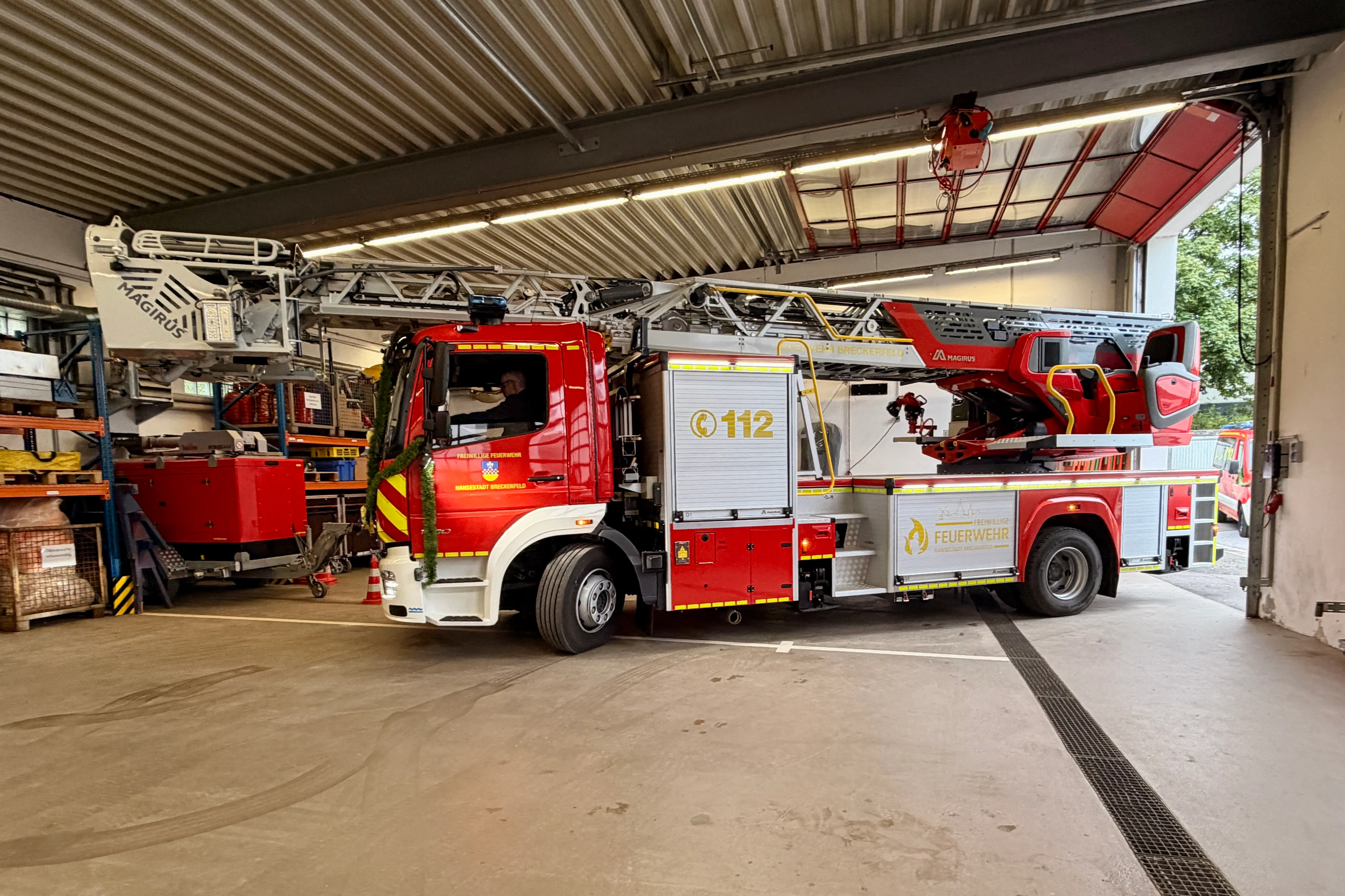 Roter Feuerwehrauto mit Drehleiter in einer hellen Fahrzeughalle, beschriftet mit „Feuerwehr“ und der Notrufnummer 112.