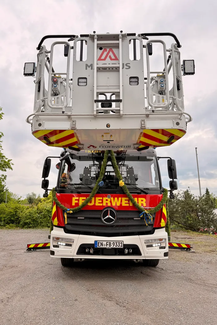 Frontale Aufnahme eines roten Feuerwehrautos der Marke Mercedes mit gelben Markierungen und einem auf dem Dach montierten Gerät. Das Fahrzeug ist mit Blumenschmuck verziert und trägt das Kennzeichen EN-FB 9331.