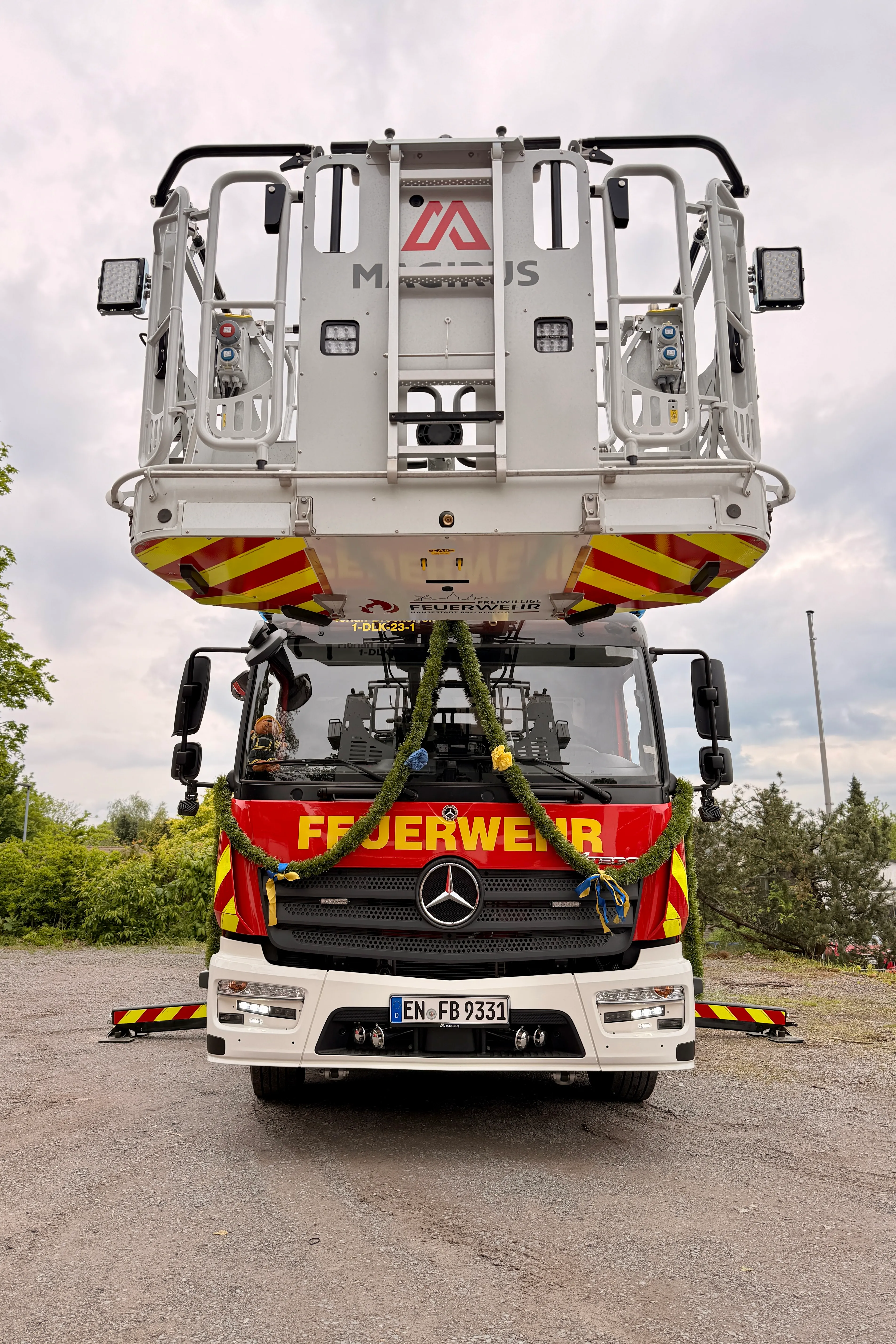 Frontale Aufnahme eines roten Feuerwehrautos der Marke Mercedes mit gelben Markierungen und einem auf dem Dach montierten Gerät. Das Fahrzeug ist mit Blumenschmuck verziert und trägt das Kennzeichen EN-FB 9331.