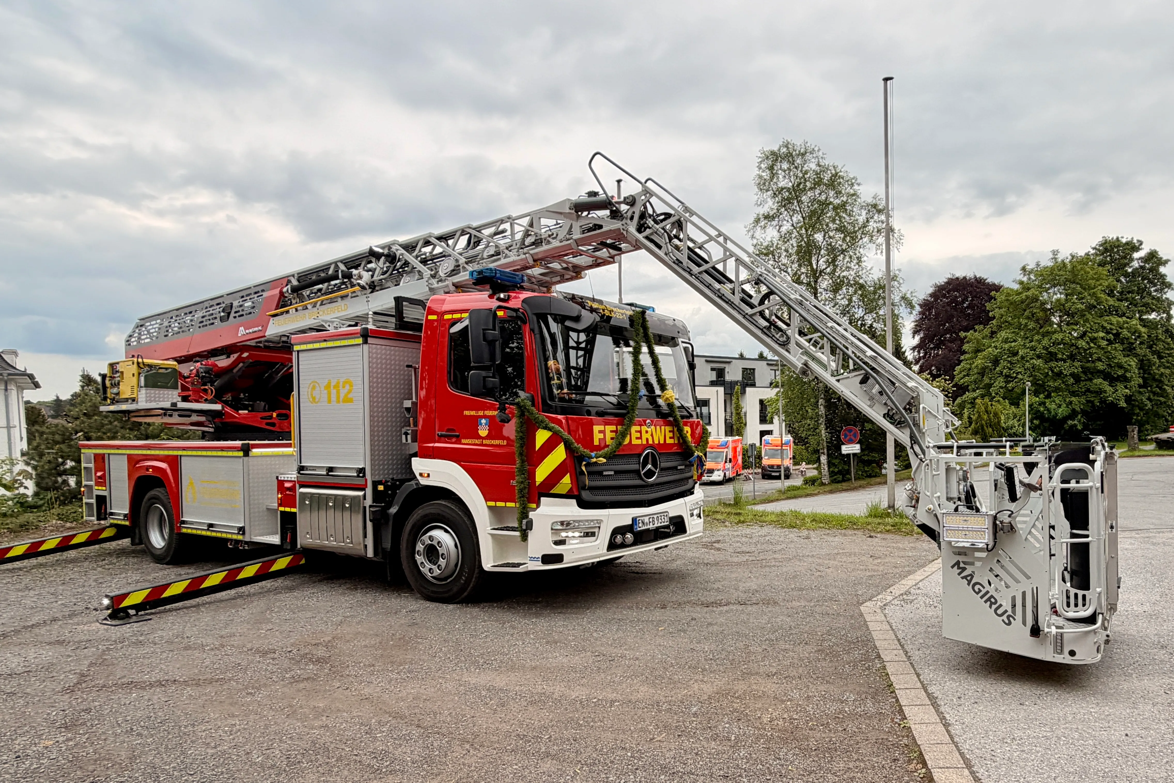 Roter Feuerwehrauto mit ausgefahrener Leiter steht auf einem Parkplatz vor Bäumen und Gebäuden. Am Fahrzeug sind Blumenketten angebracht.