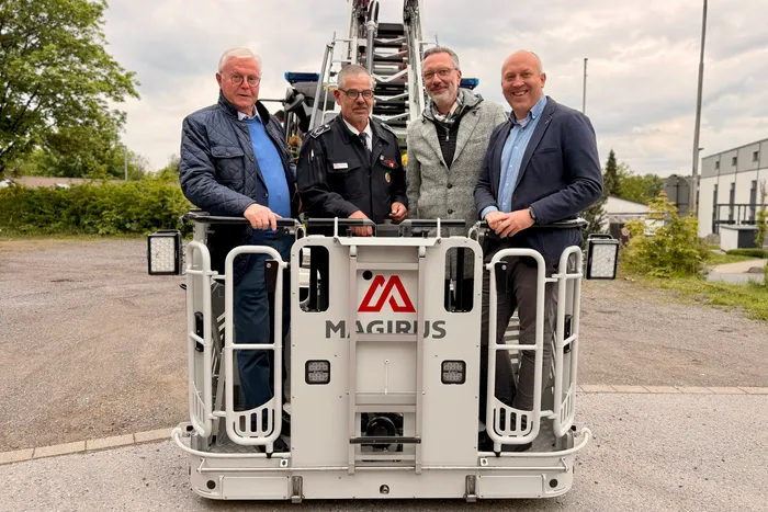 Vier Männer stehen vor einem weißen Feuerwehrauto mit dem Aufdruck „Magirus“. Im Hintergrund ein Gebäude und Bäume.