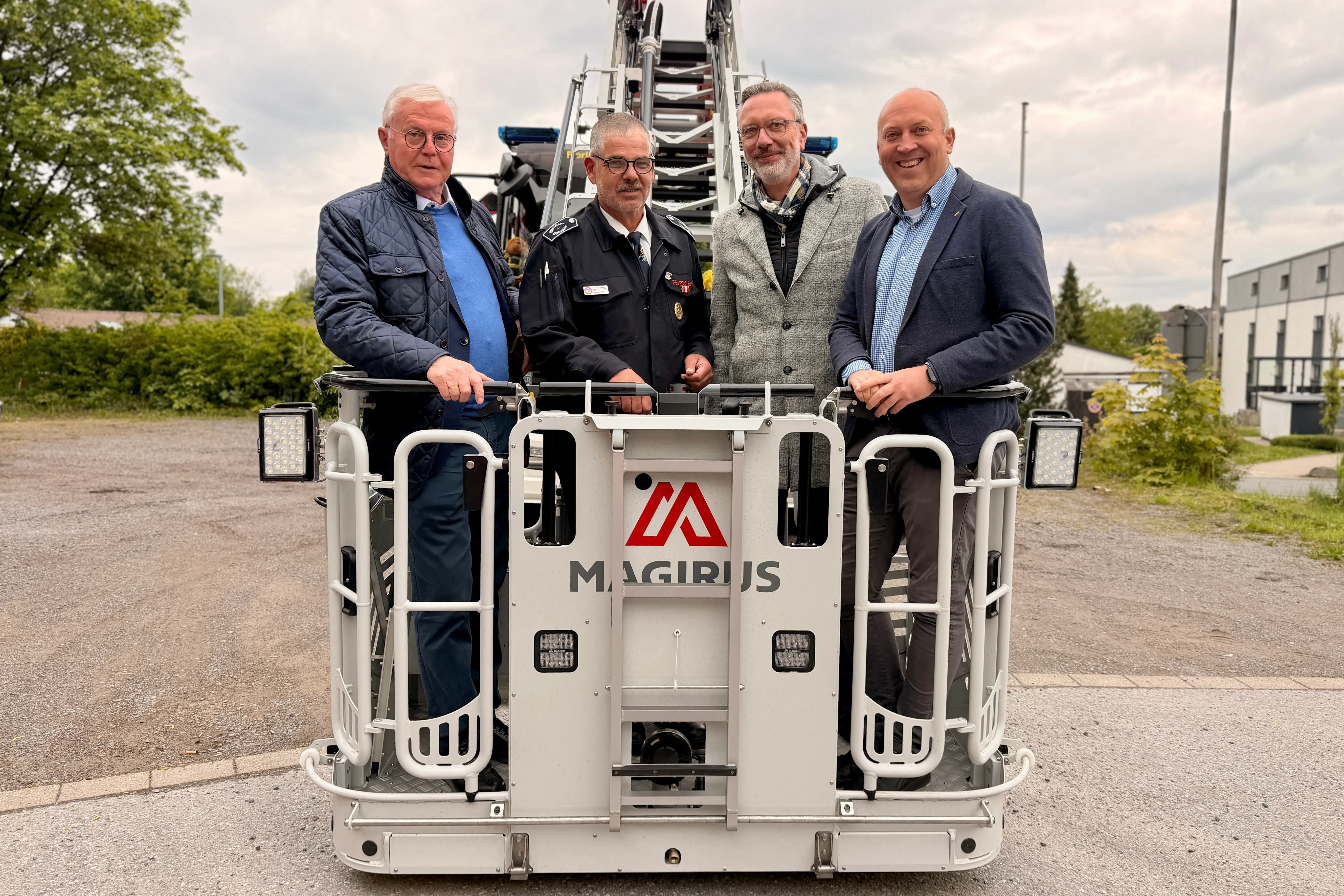 Vier Männer stehen vor einem weißen Feuerwehrauto mit dem Aufdruck „Magirus“. Im Hintergrund ein Gebäude und Bäume.