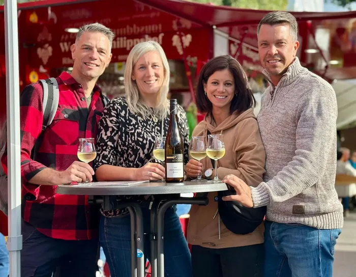 Vier lächelnde Menschen stehen an einem Tisch im Freien, jeder hält ein Glas Wein. Eine Flasche Wein steht in der Mitte des Tisches. Im Hintergrund ist ein Weinfeststand mit Dekorationen zu sehen.