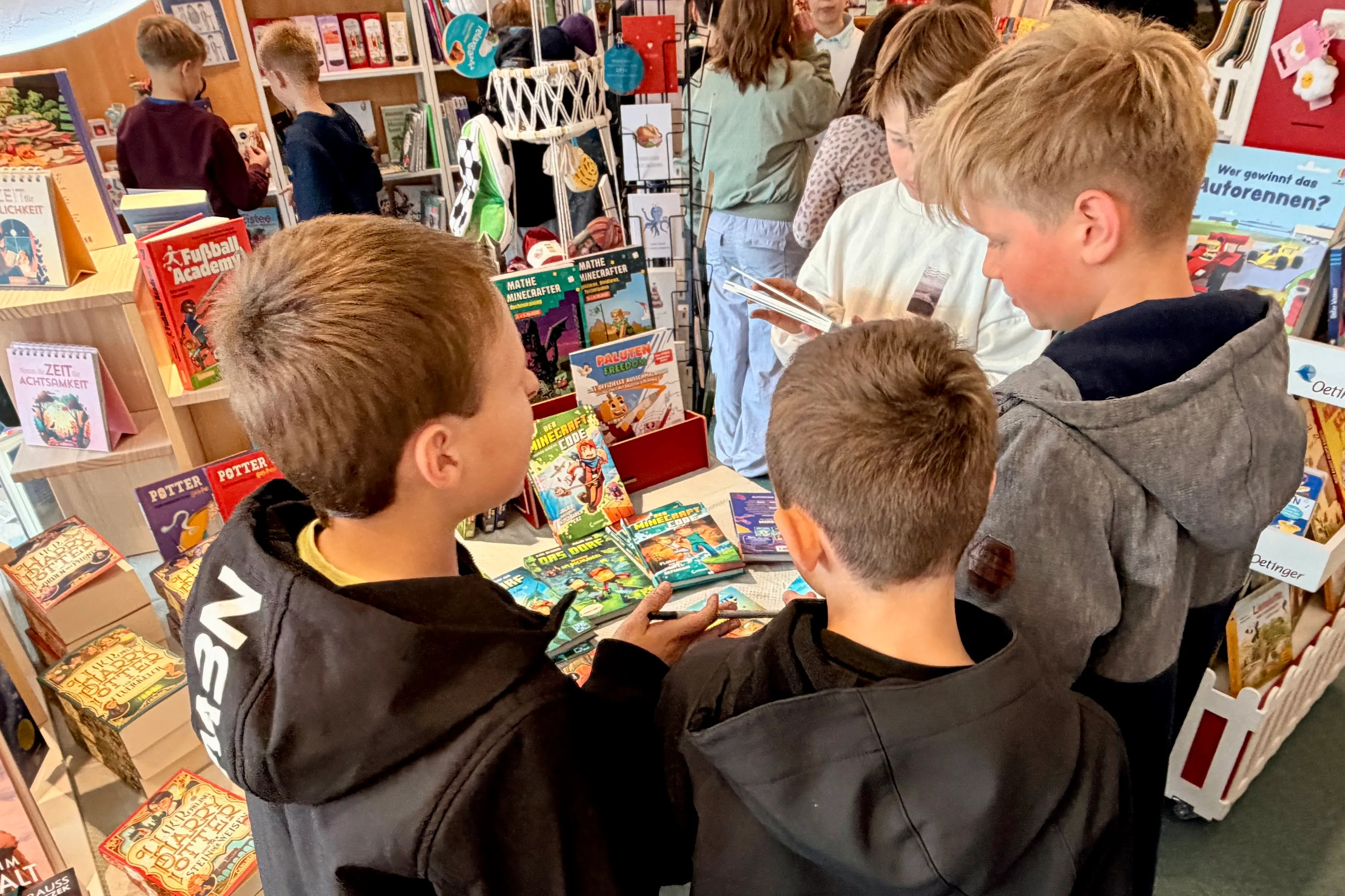 Mehrere Schüler*innen betrachten Bücher an einem Tisch in einer Buchhandlung, umgeben von weiteren Besuchern und Regalen voller Bücher.