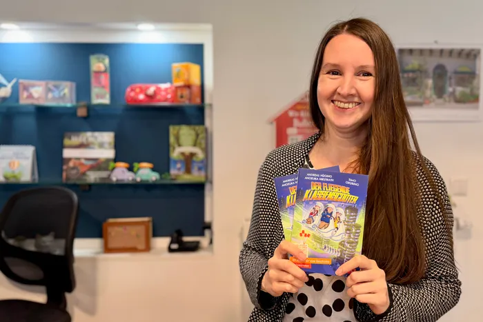 Frau mit braunen Haaren lächelt in einer Buchhandlung und hält ein Kinderbuch mit dem Titel „Der fliegende Klassenzimmerleiter“ in der Hand.