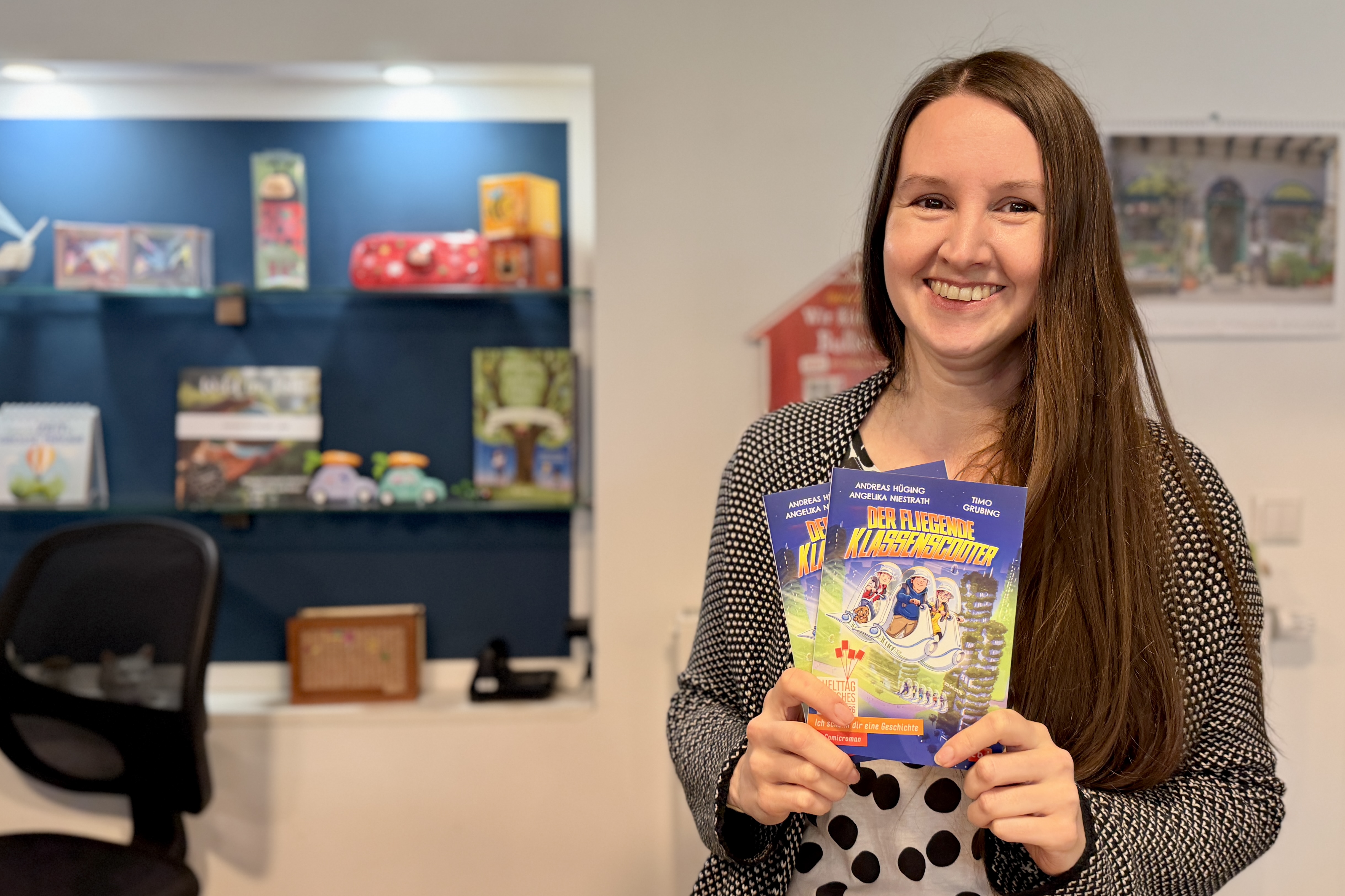 Frau mit braunen Haaren lächelt in einer Buchhandlung und hält ein Kinderbuch mit dem Titel „Der fliegende Klassenzimmerleiter“ in der Hand.