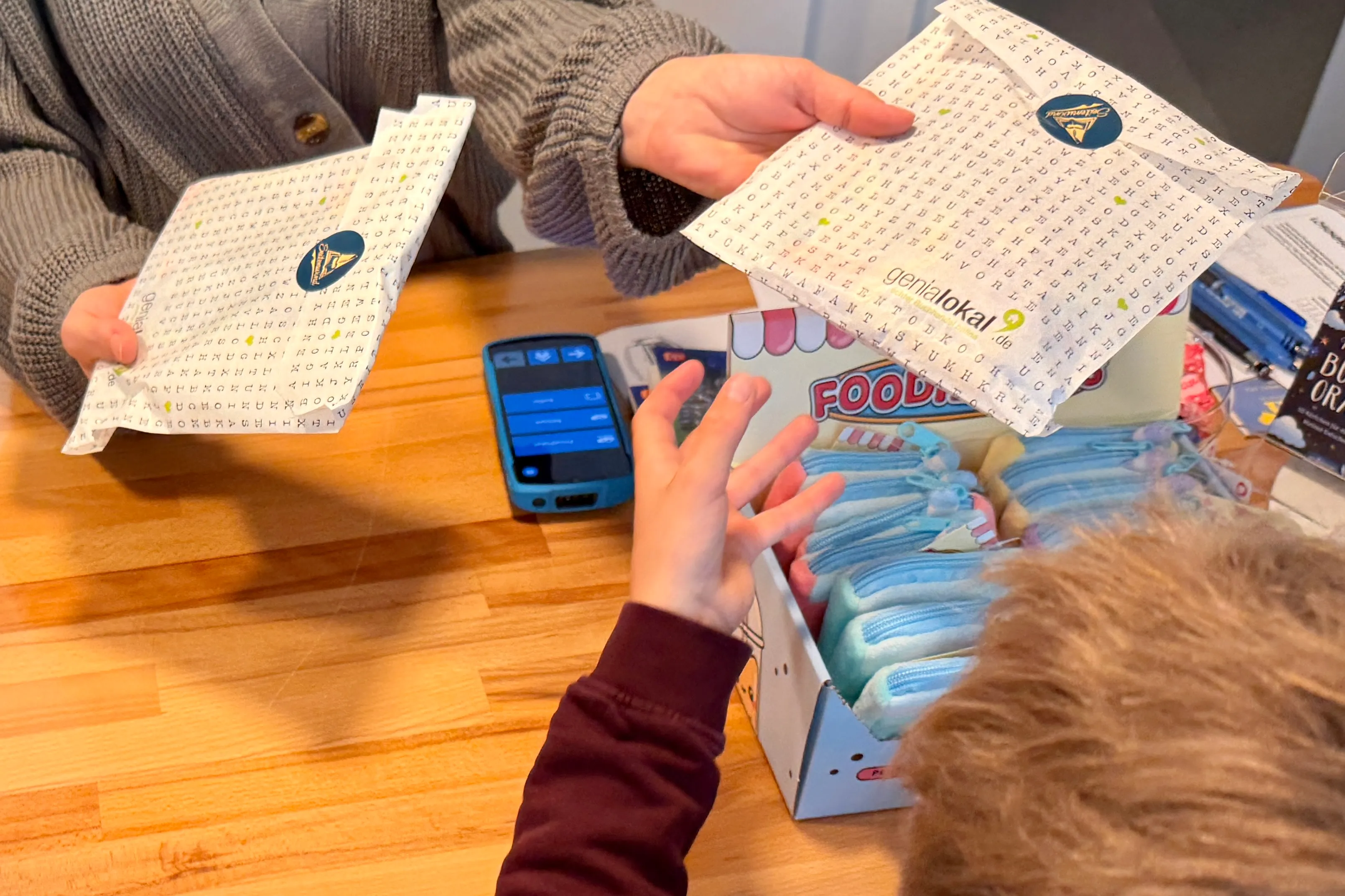 Zwei Hände halten jeweils ein quadratisches Papier mit Buchstabenmustern, während eine dritte Hand ein Smartphone darüber hält. Im Hintergrund sind weitere Gegenstände wie Bücher und eine Verpackung zu sehen.