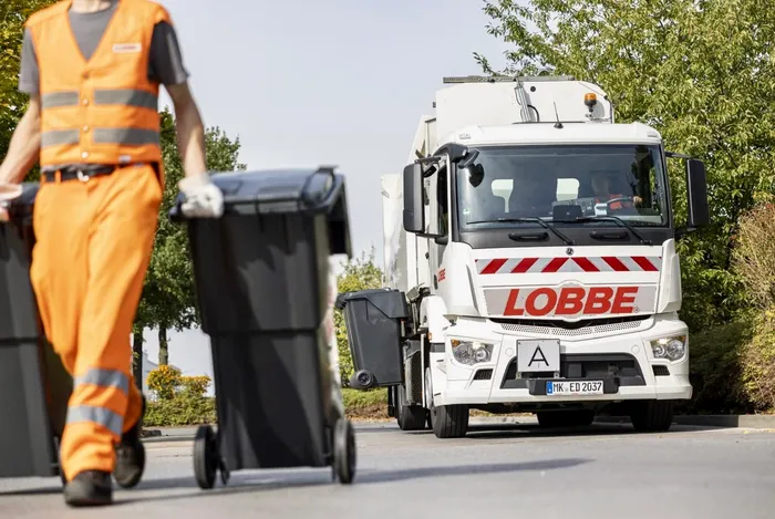 Ein weißer Müllwagen mit dem Schriftzug „LOBBE“ fährt auf einer Straße, während ein Müllwerker in orangefarbener Arbeitskleidung Mülltonnen schiebt.