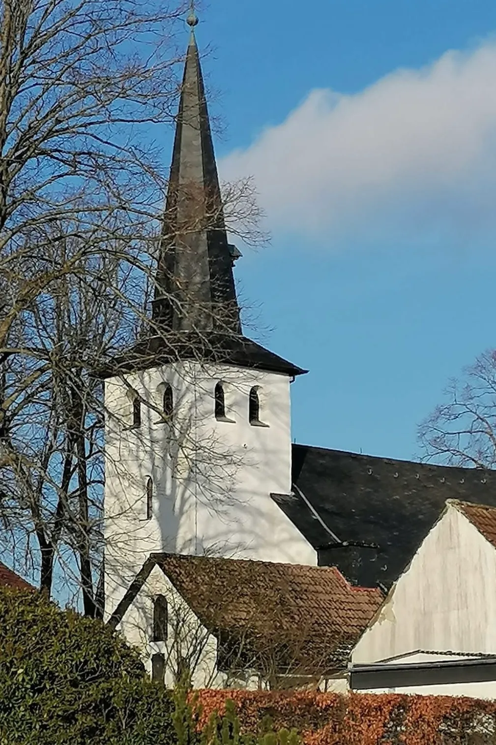Weißer Kirchturm mit steilem, dunklen Dach und kleinem goldenen Kreuz an der Spitze, Teil eines größeren Kirchengebäudes mit roten Ziegeldächern.