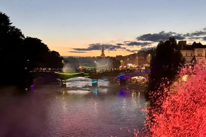 Beleuchtete Brücke mit Menschen im Abendlicht, im Hintergrund Gebäude und ein Kirchturm.