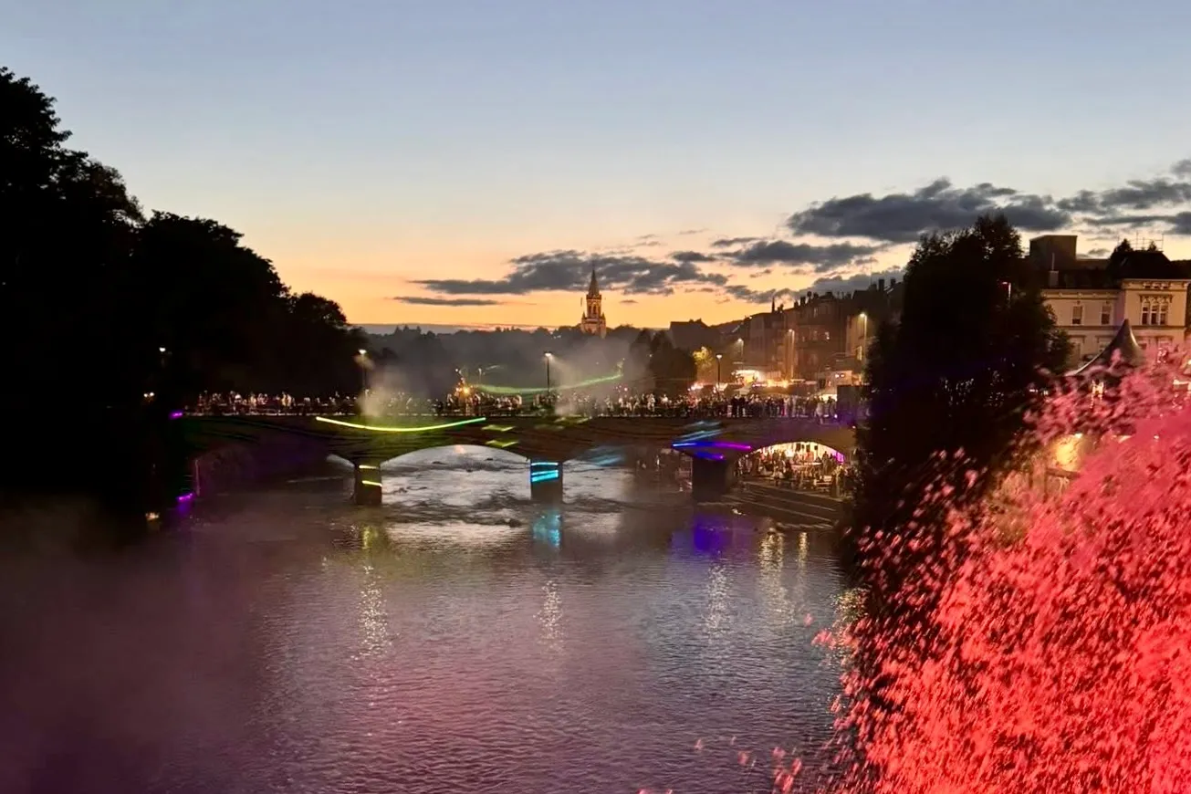 Beleuchtete Brücke mit Menschen im Abendlicht, im Hintergrund Gebäude und ein Kirchturm.