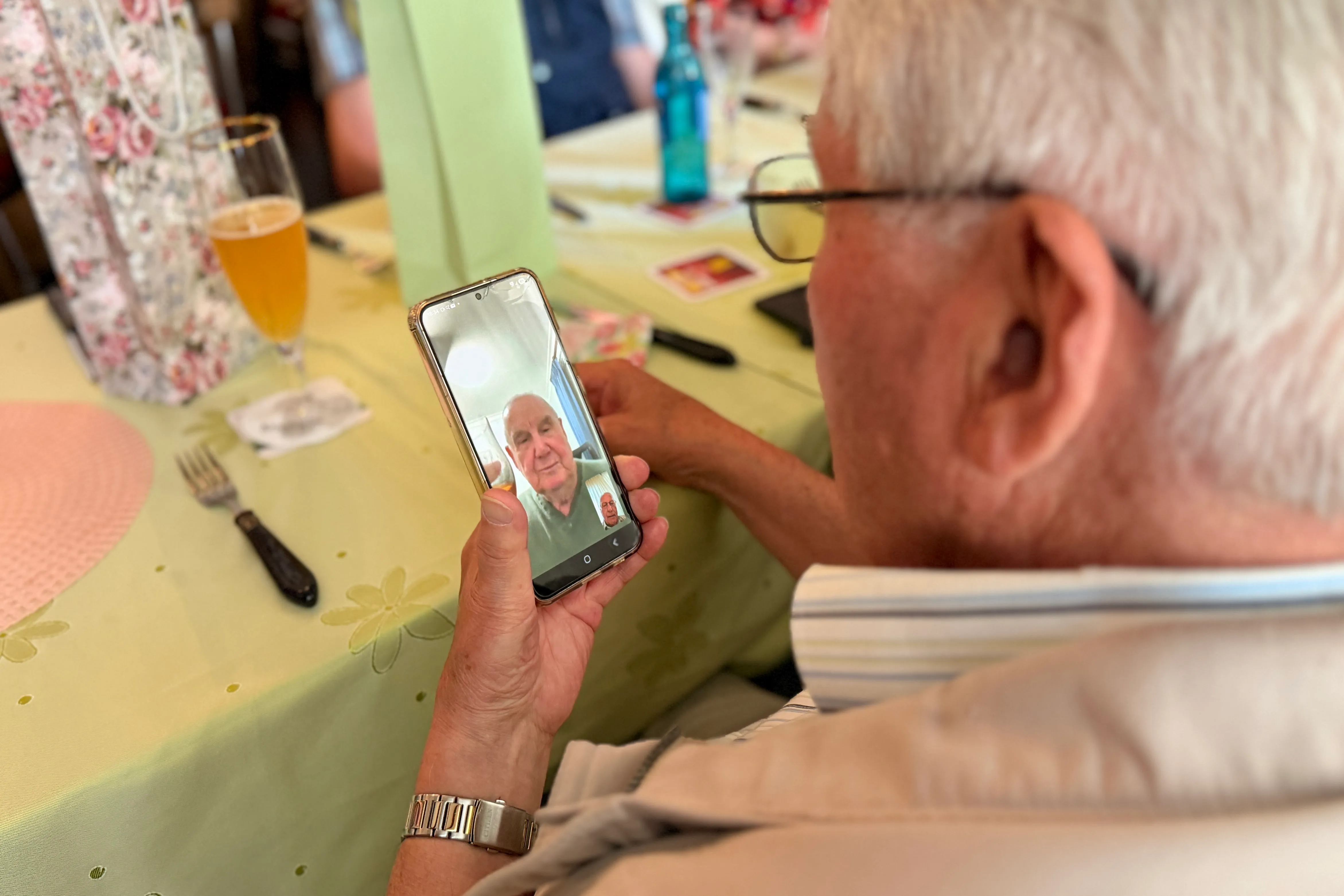 Ein älterer Mann mit Brille hält ein Smartphone vor sich, auf dem ein weiterer älterer Mann im Videochat zu sehen ist. Im Hintergrund ein gedeckter Tisch mit Getränken und Besteck.