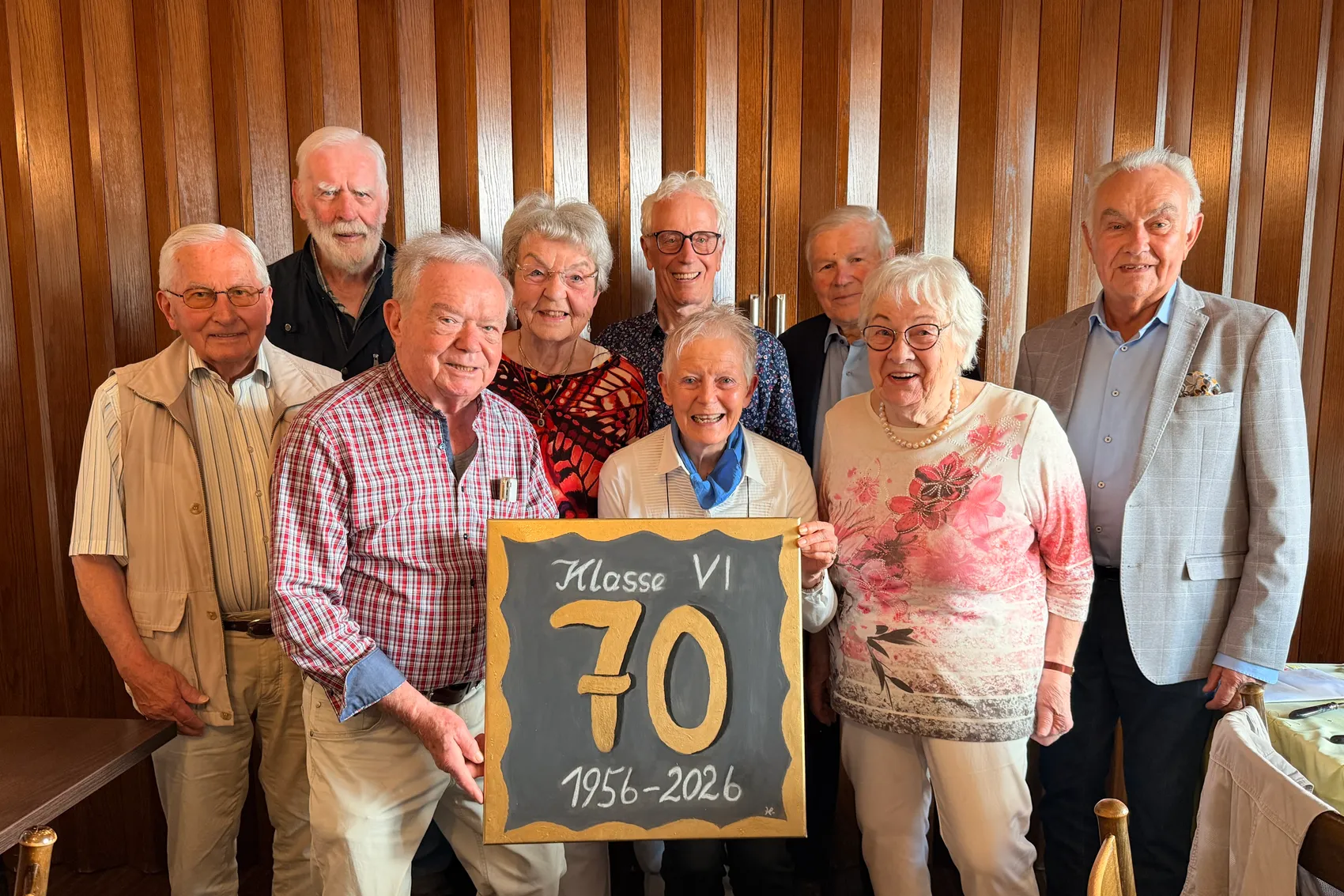 Neun ältere Menschen stehen lächelnd zusammen, eine Frau hält eine Tafel mit der Aufschrift „Klasse VI 70 1956-2026“.