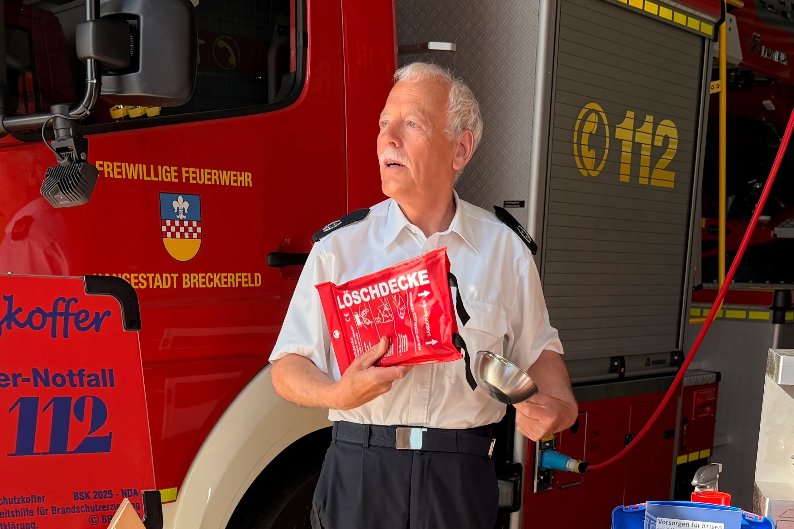 Ein älterer Mann in Uniform der Freiwilligen Feuerwehr hält eine Brandschutzdecke in der Hand. Im Hintergrund ist ein rotes Feuerwehrauto zu sehen.