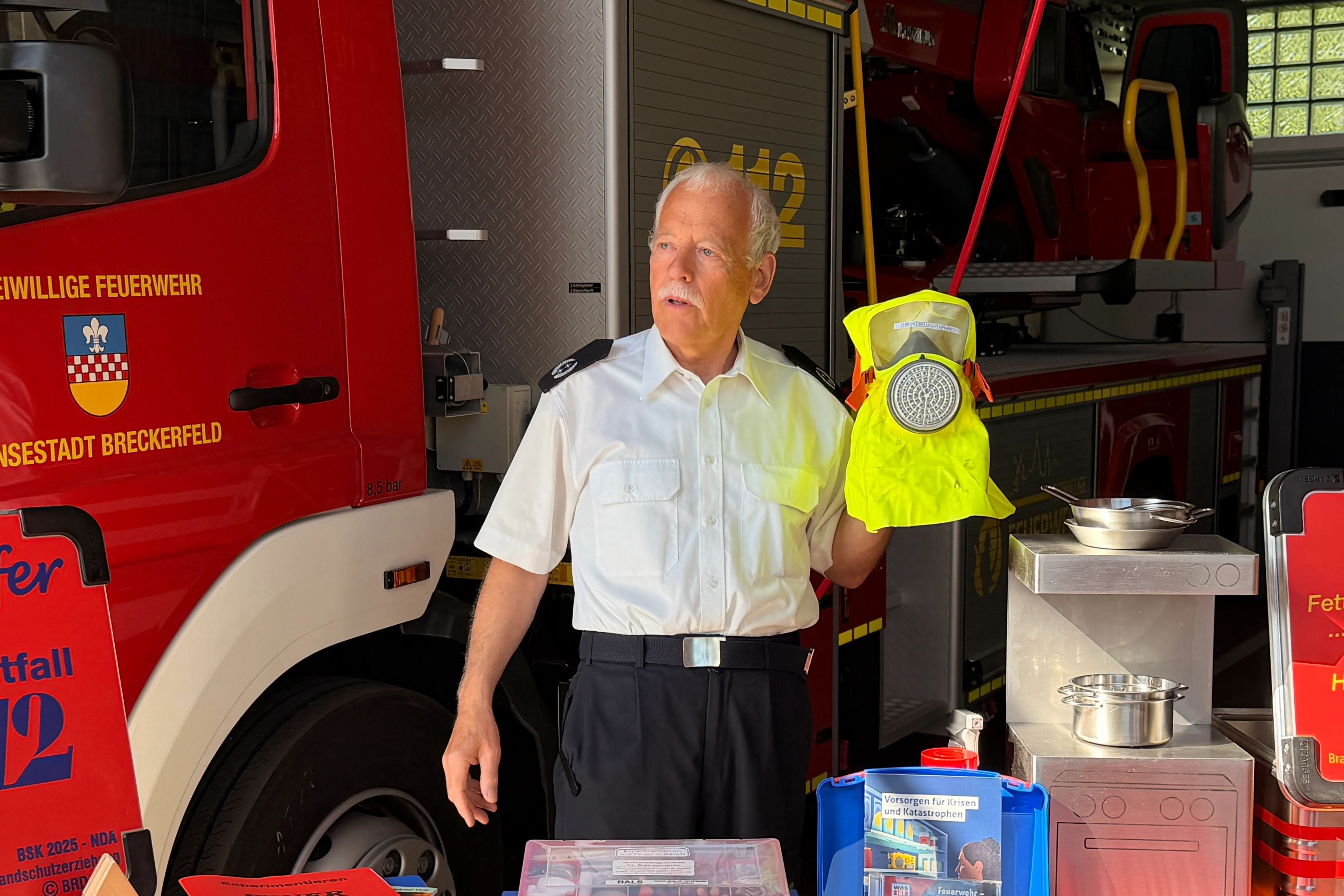 Ein älterer Feuerwehrmann in Uniform spricht in eine Handmikrofon vor einem roten Feuerwehrauto und Ausrüstung.