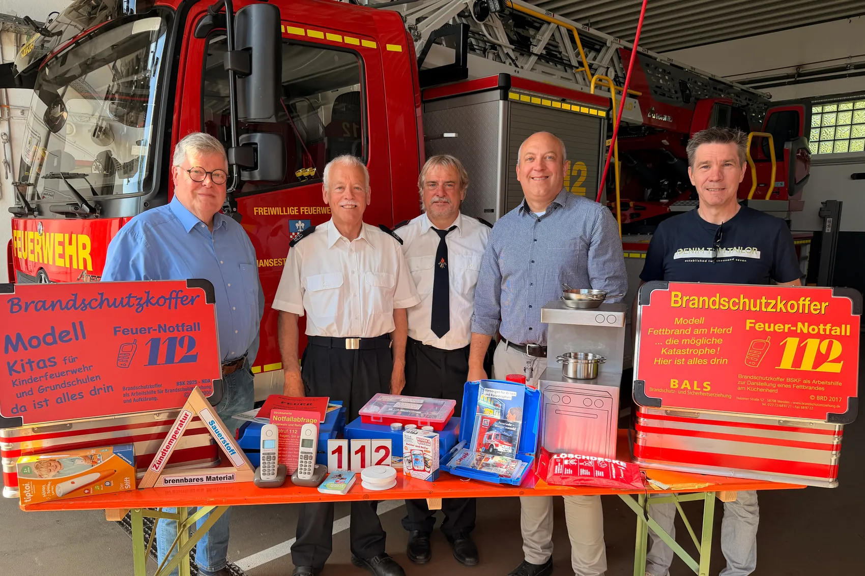 Fünf Personen stehen vor einem Tisch mit Brandschutzmaterialien und einem Feuerwehrauto. Die drei Kuratoriumsmitglieder der Bürgerstiftung überreichten Lehrmaterial an zwei Brandschutzerzieher.