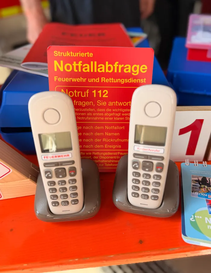 Zwei rote Spielzeugtelefone stehen vor einem roten Schild mit der Aufschrift „Notruf 112“. Sie dienen zum Üben des richtigen Verhaltens bei einem Notruf im Rahmen der Brandschutzerziehung.