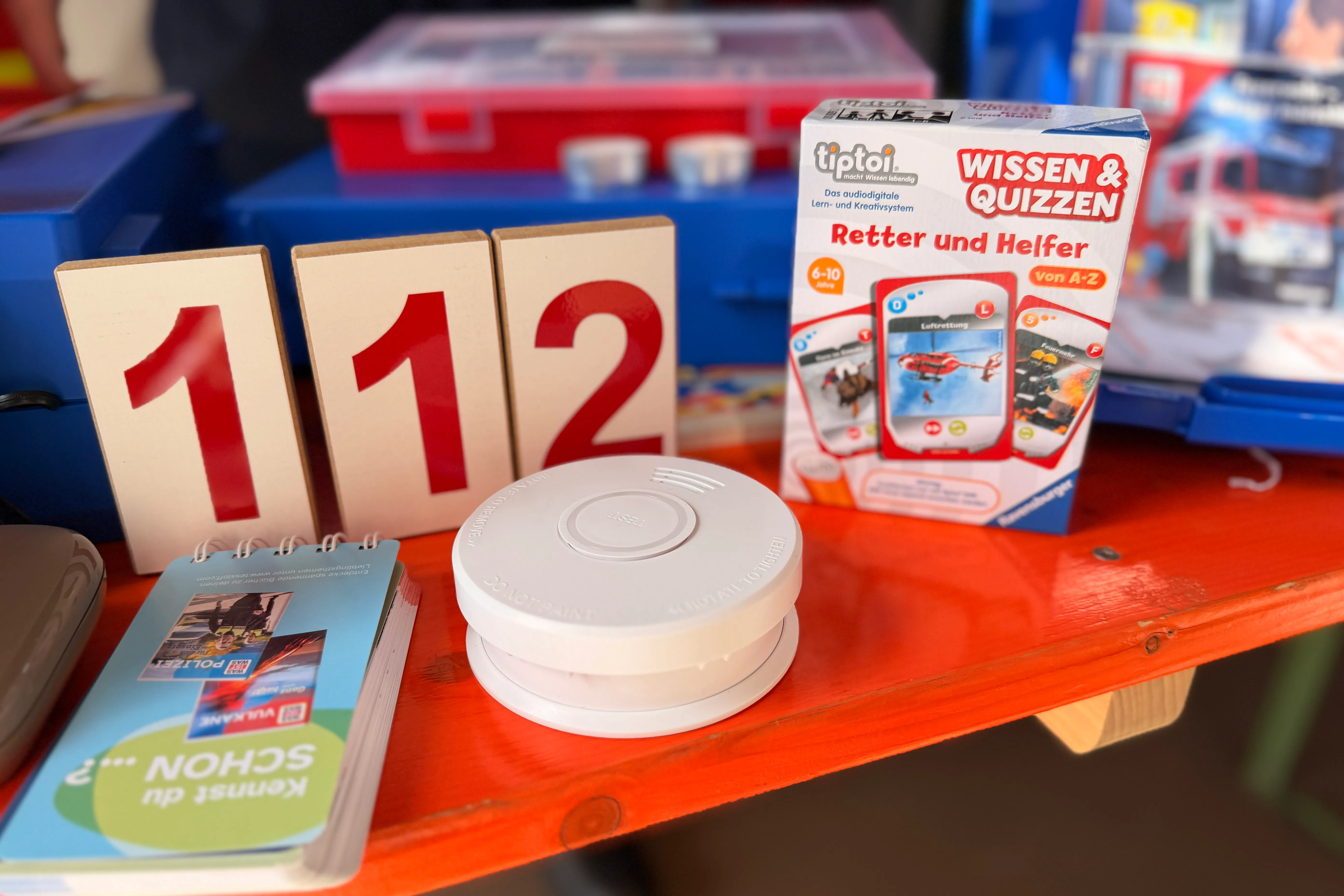 Notrufnummer 112, Rauchmelder und Lernmaterialien für Kinder stehen auf einem Holztisch.
