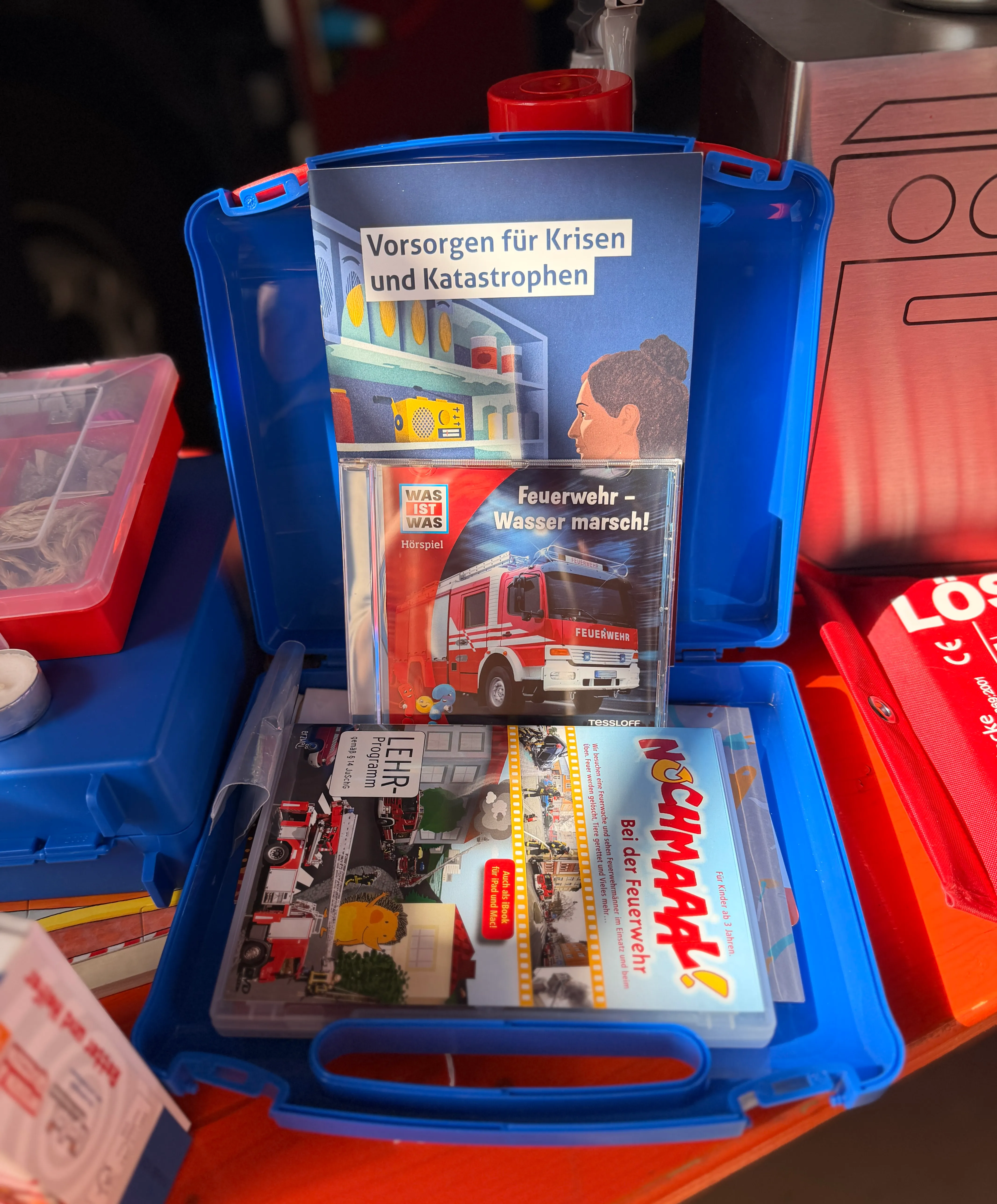 Offener blauer Koffer mit kindgerechten Materialien zum Thema Feuerwehr und Katastrophenschutz, darunter Bücher, eine Spielzeugfigur und eine rote Schachtel.