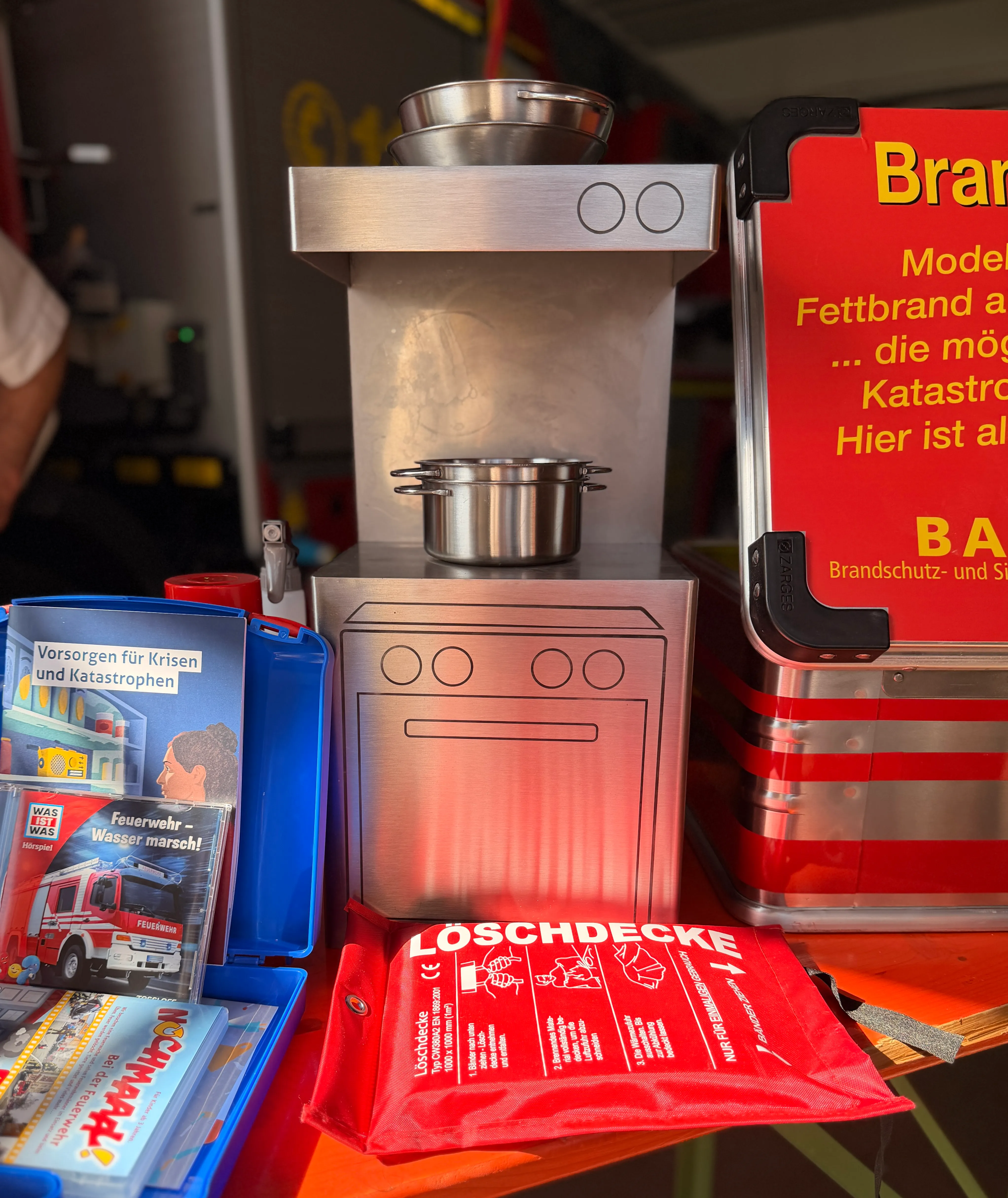 Eine Edelstahl-Fettbrandlöschanlage mit einem Topf darauf steht vor Büchern und einer blauen Kiste mit dem Aufdruck „Feuerwehr – Wasser marsch!“ und einer roten Löschdecke.