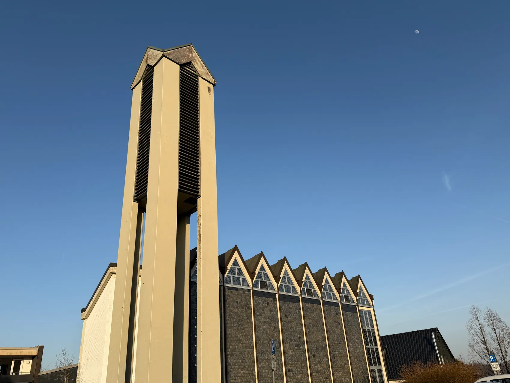Moderner Kirchenbau mit markantem, hoch aufragendem Glockenturm und zackigem Giebeldach unter blauem Himmel.