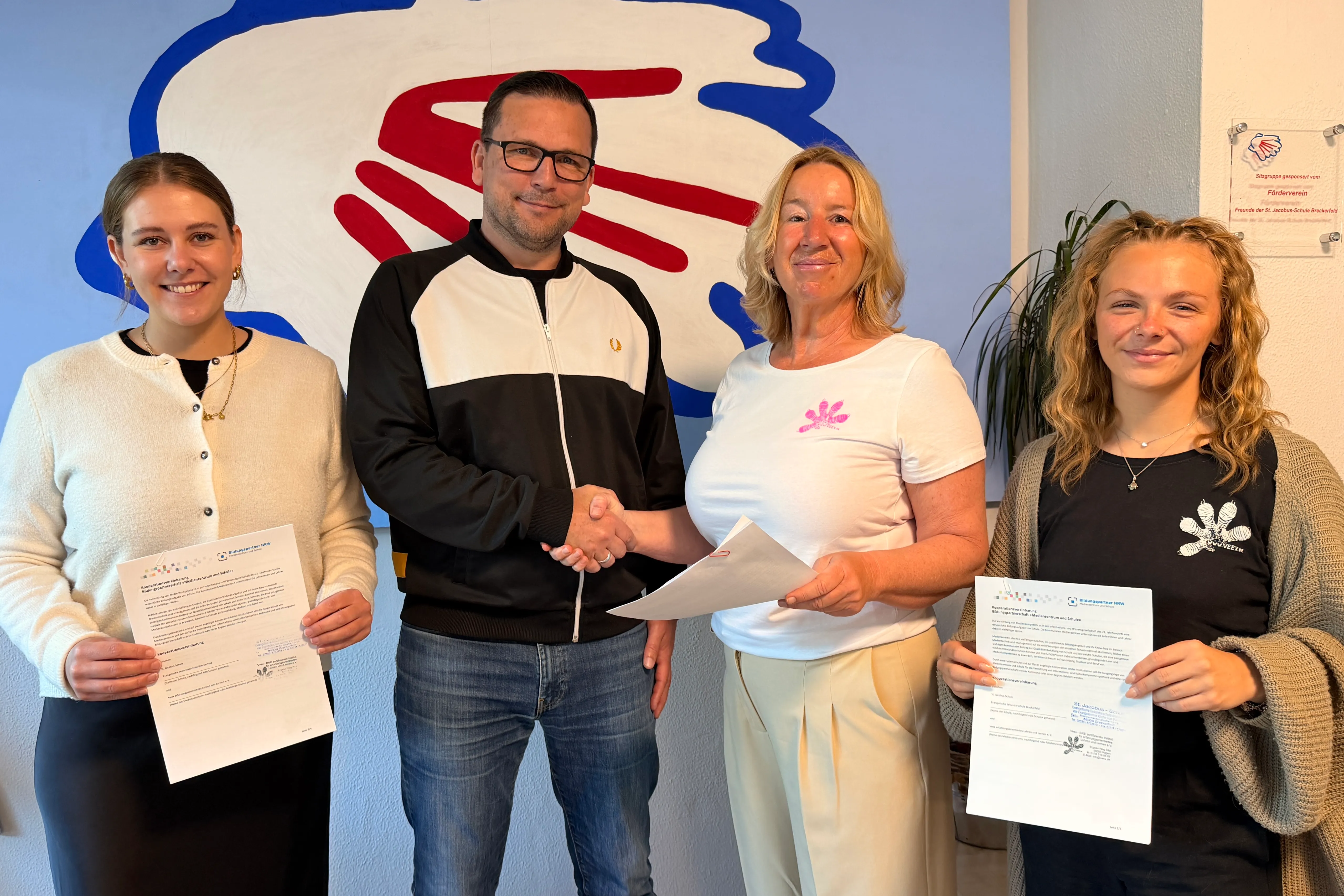 Schulleiter Dirk Bollwahn gibt Schulsozialarbeiterin Pascalina Tophoven, Veex-Institutsleiterin Andrea Bock-Dombrowski und Junia Dröge vom Veex-Team Urkunden. Im Hintergrund eine stilisierte Darstellung von Händen.