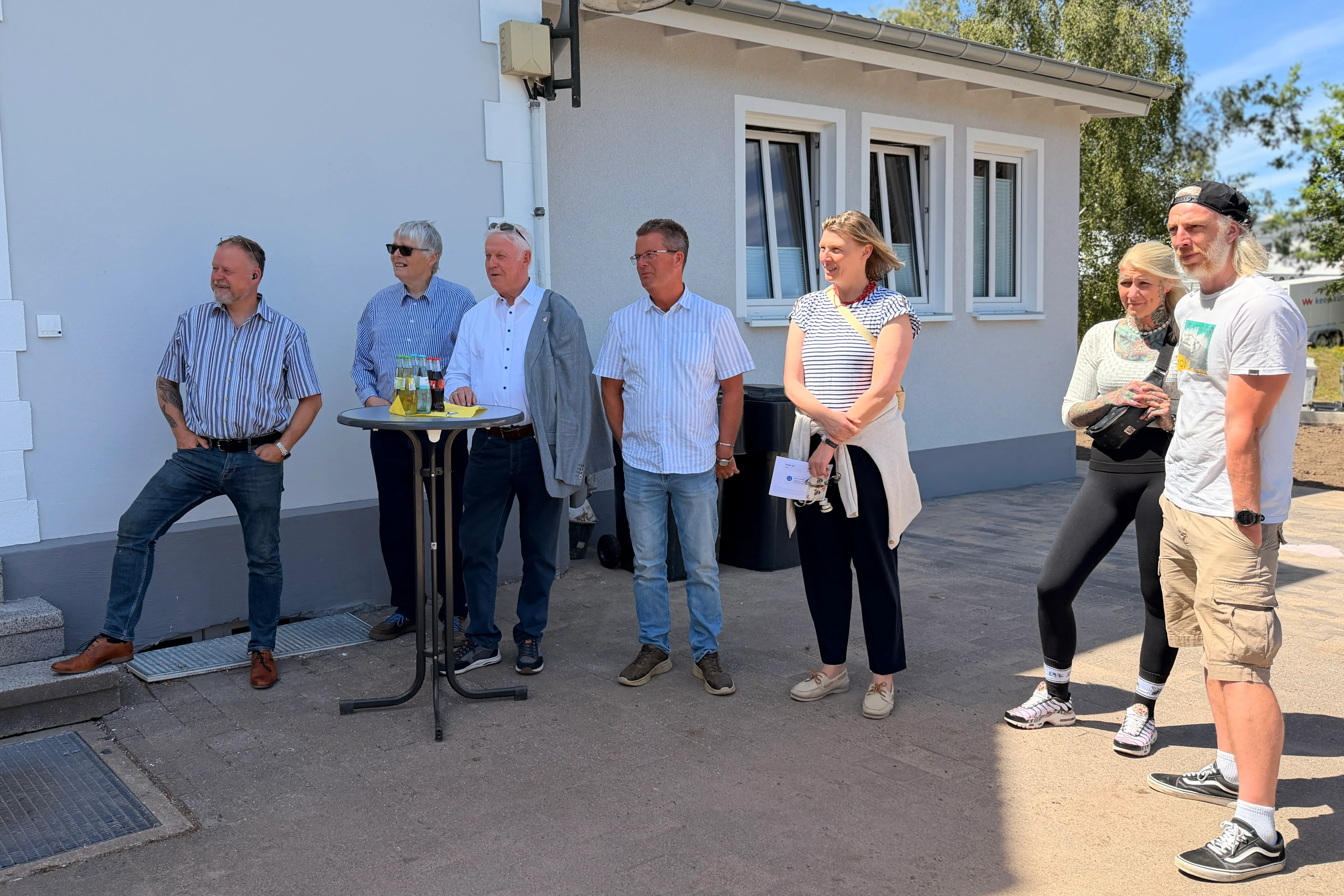 Eine Gruppe von sechs Personen steht vor einem hellen Gebäude mit Fenstern. Sie scheinen an einer Eröffnungsfeier teilzunehmen, möglicherweise im Zusammenhang mit einem Jugendzentrum.