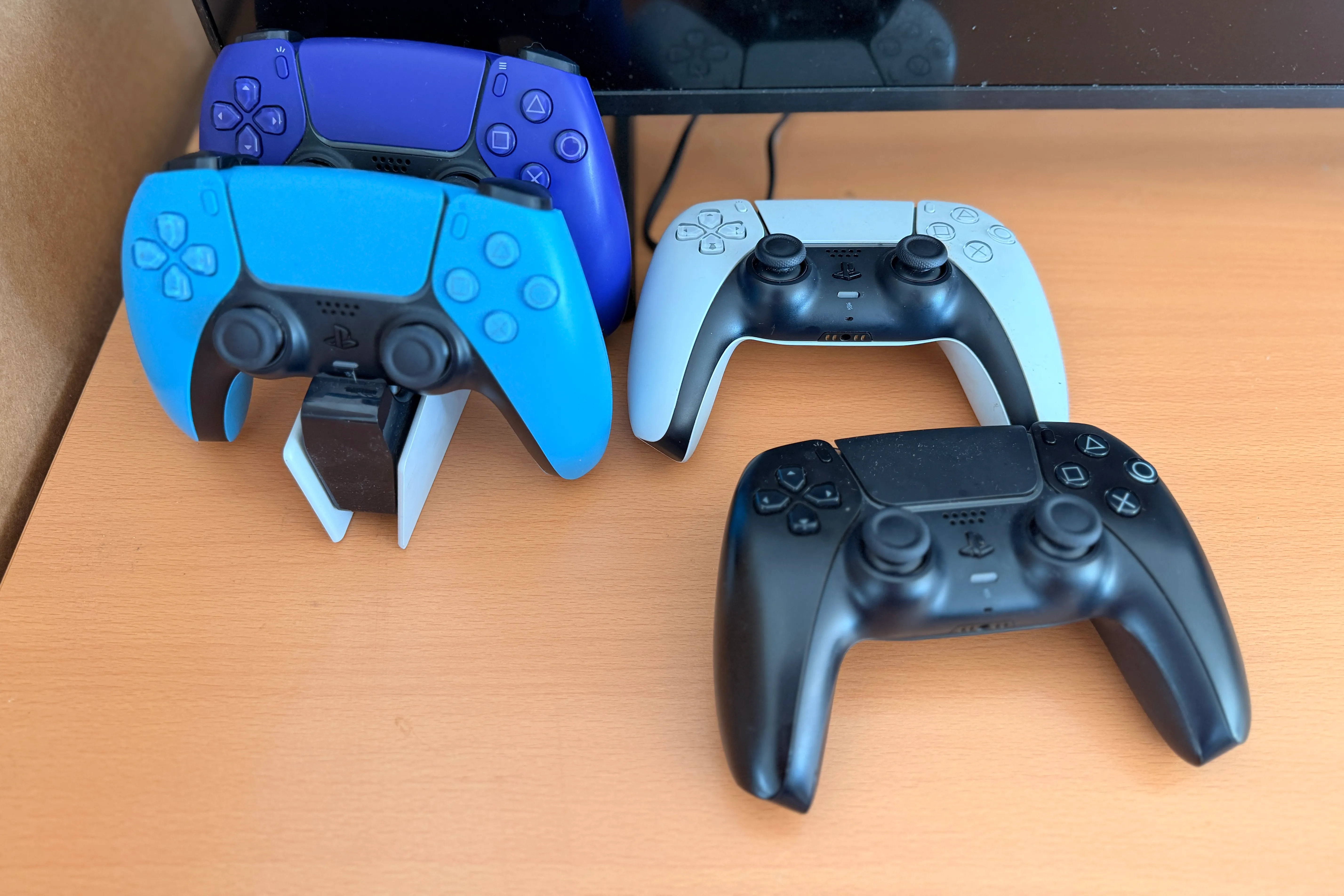 Drei verschiedene Playstation-Controller in Blau, Weiß und Schwarz liegen auf einer hellen Holzoberfläche.