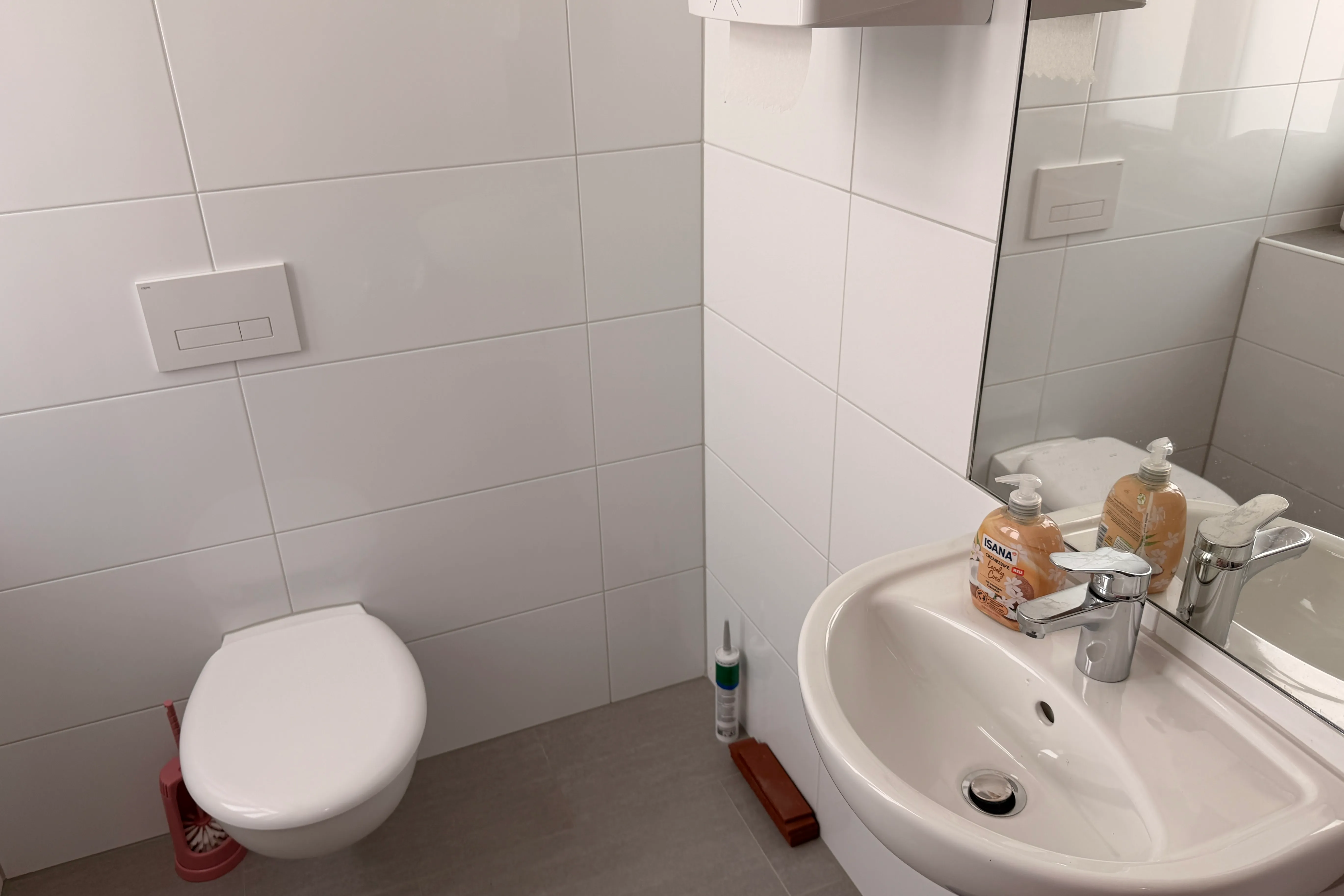 Helles Badezimmer mit weiß gefliesten Wänden, einer Toilette und einem Waschbecken mit Armatur und Seifenspendern.