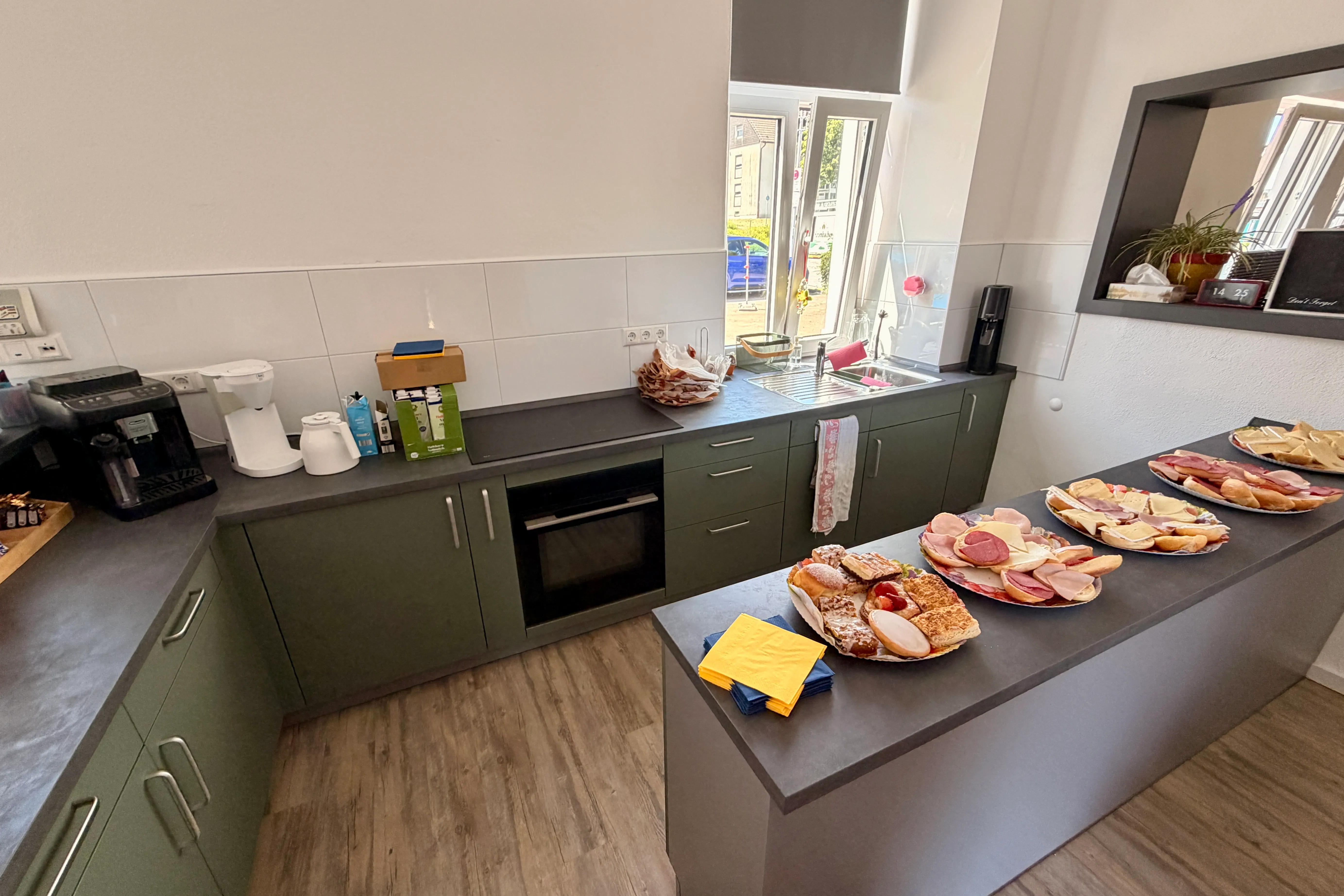 Eine moderne Küche mit grauen Schränken und Arbeitsplatten, auf der eine Auswahl an Aufschnitt, Brot und Gebäck präsentiert wird. Im Hintergrund ein Fenster mit Blick auf einen Garten.