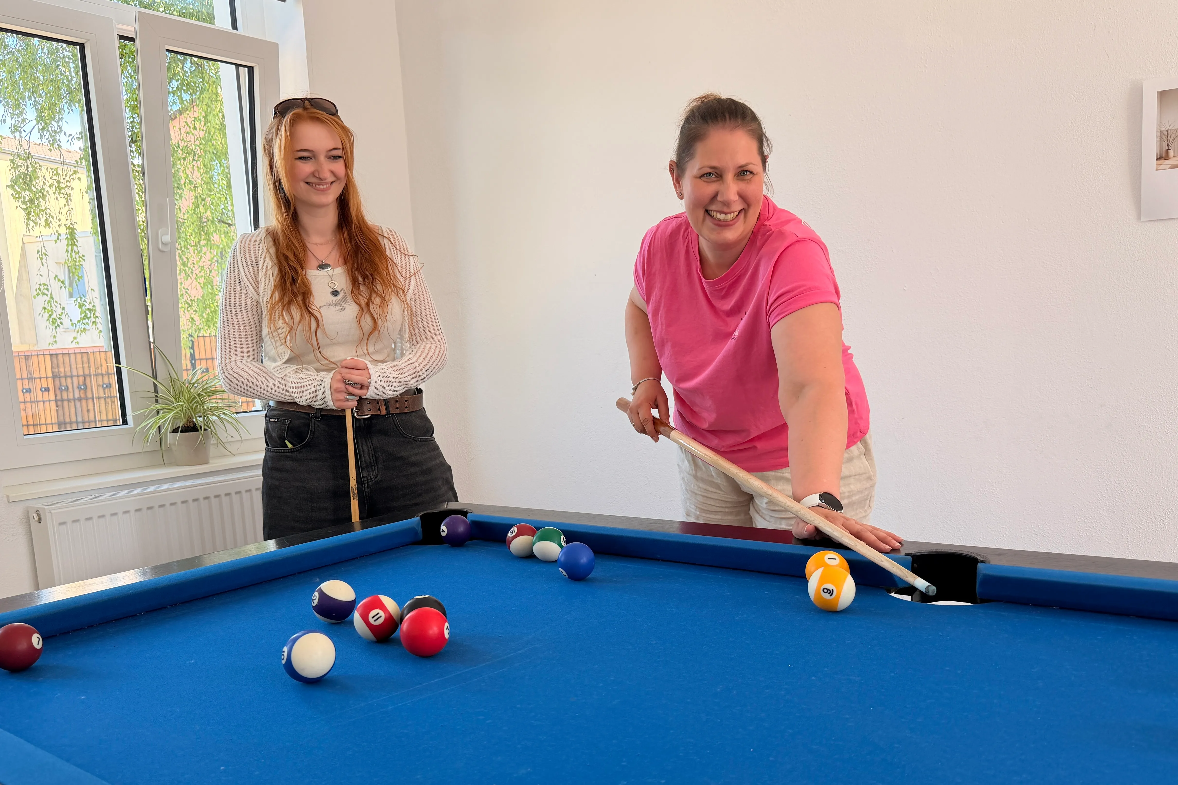 Zwei lächelnde Frauen spielen Billard in einem hellen Raum des Jugendzentrums. Im Hintergrund ein Fenster mit Blick auf Bäume.