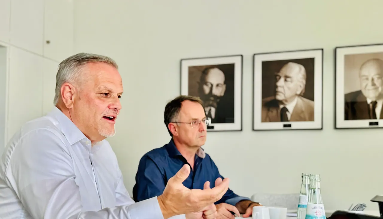 Andreas Engelhardt spricht gestikulierend mit Dr. Walter Stadlbauer an einem Konferenztisch, im Hintergrund Porträts der Gründerfamilie Fuchs.