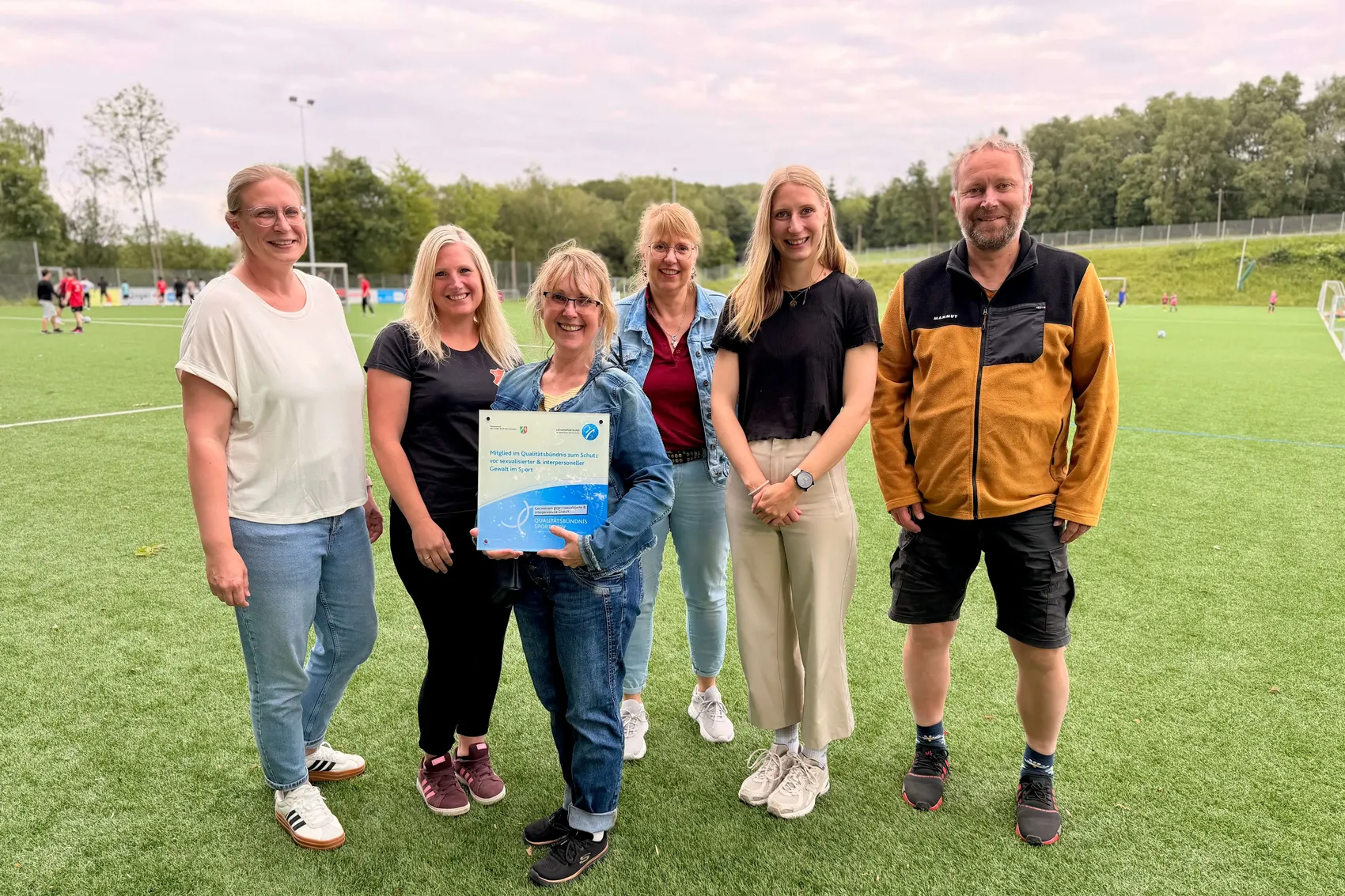 Fünf Frauen und ein Mann stehen lächelnd auf einem Sportplatz und halten eine Urkunde. Die Urkunde bestätigt die Aufnahme des SC Zurstrasse in ein Qualitätsbündnis zum Schutz vor Gewalt im Sport.