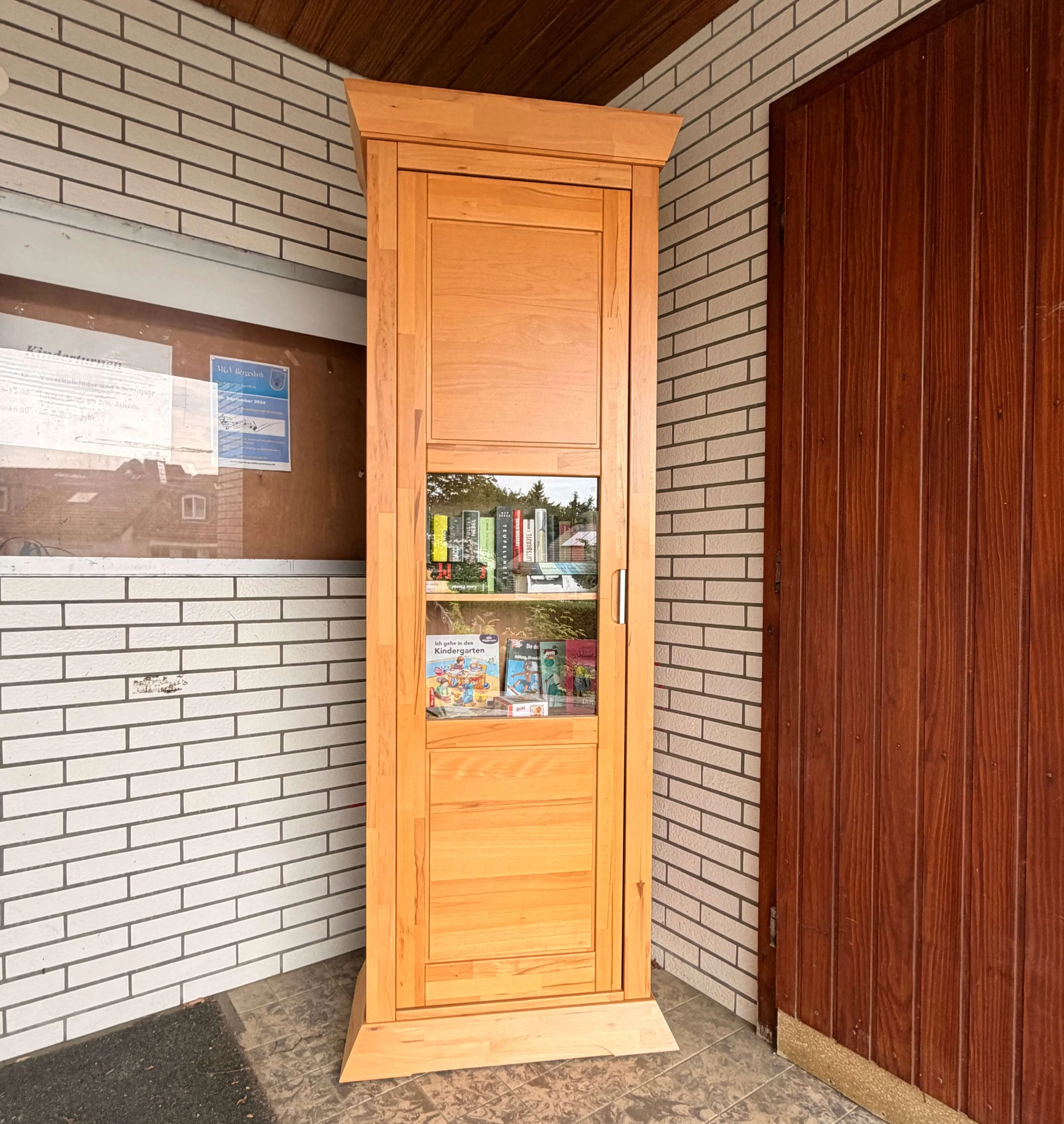Holz-Bücherschrank mit Glasfront steht vor einer gemauerten Fassade. Bücher und Spielzeug sind im Inneren zu sehen.