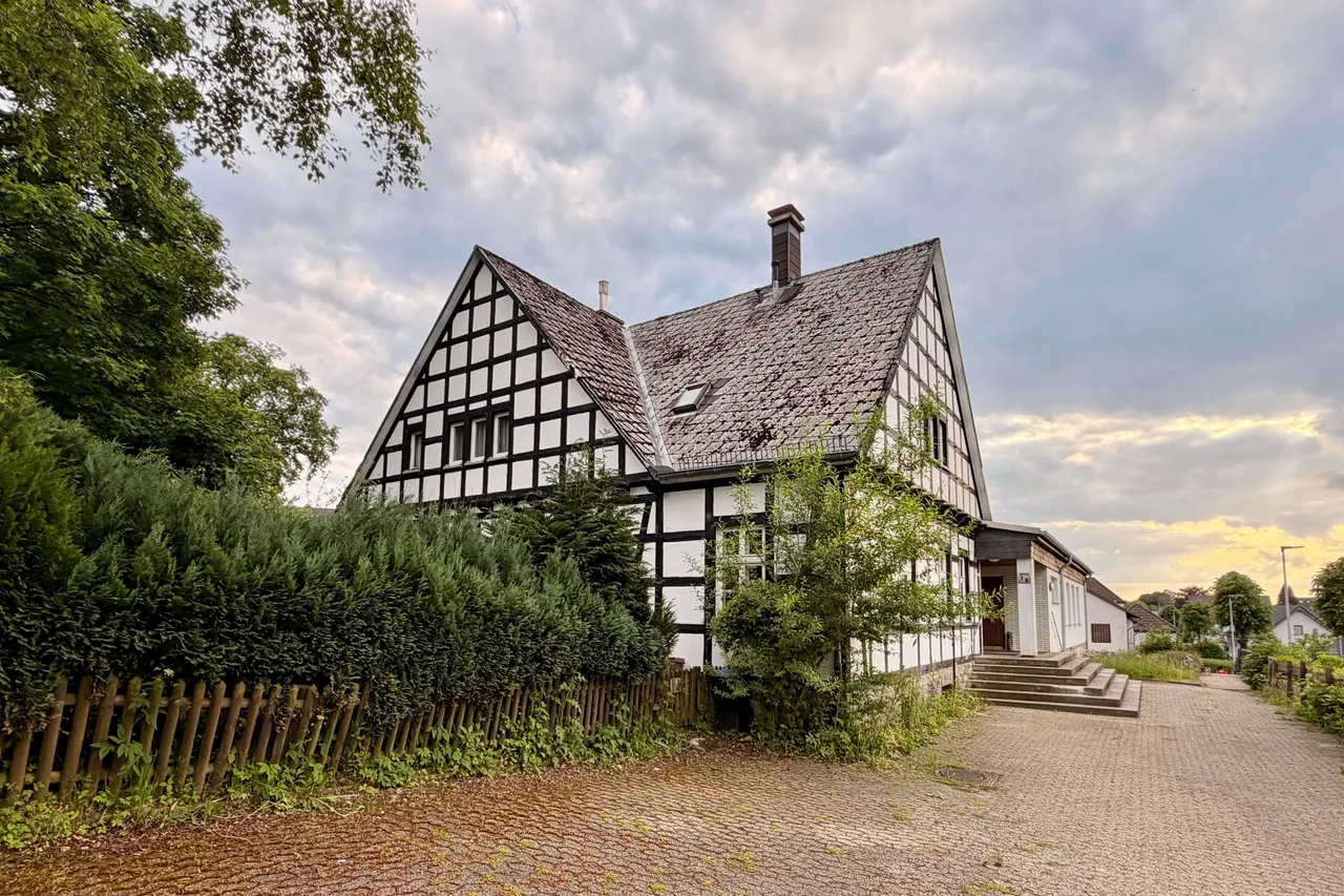 Fachwerkhaus mit steilem Dach und Schornstein an einer Straße, umgeben von Grün und einem Holzzaun.