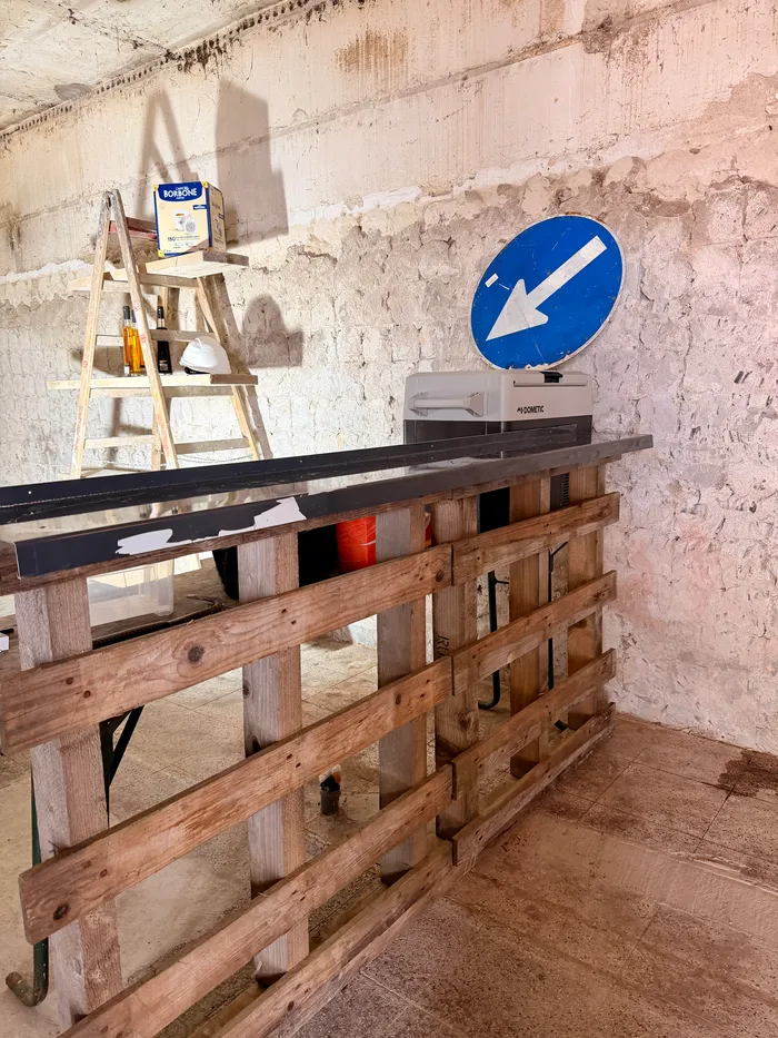 Ein hölzerner Steg führt vor einer hellen, unregelmäßigen Steinwand in einen Raum. Auf der Wand steht ein blaues Verkehrsschild mit Pfeil und darüber eine schwarze Maschine mit der Aufschrift 