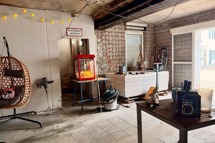 Ein Raum mit unfertigen Wänden und Decken, der sich in einem Umbau befindet. Links ein Korbsessel mit einer Katze darauf, in der Mitte ein Popcorn-Automat und rechts Werkzeuge und eine Werkbank. Im Hintergrund eine geschlossene Tür und ein Fenster.