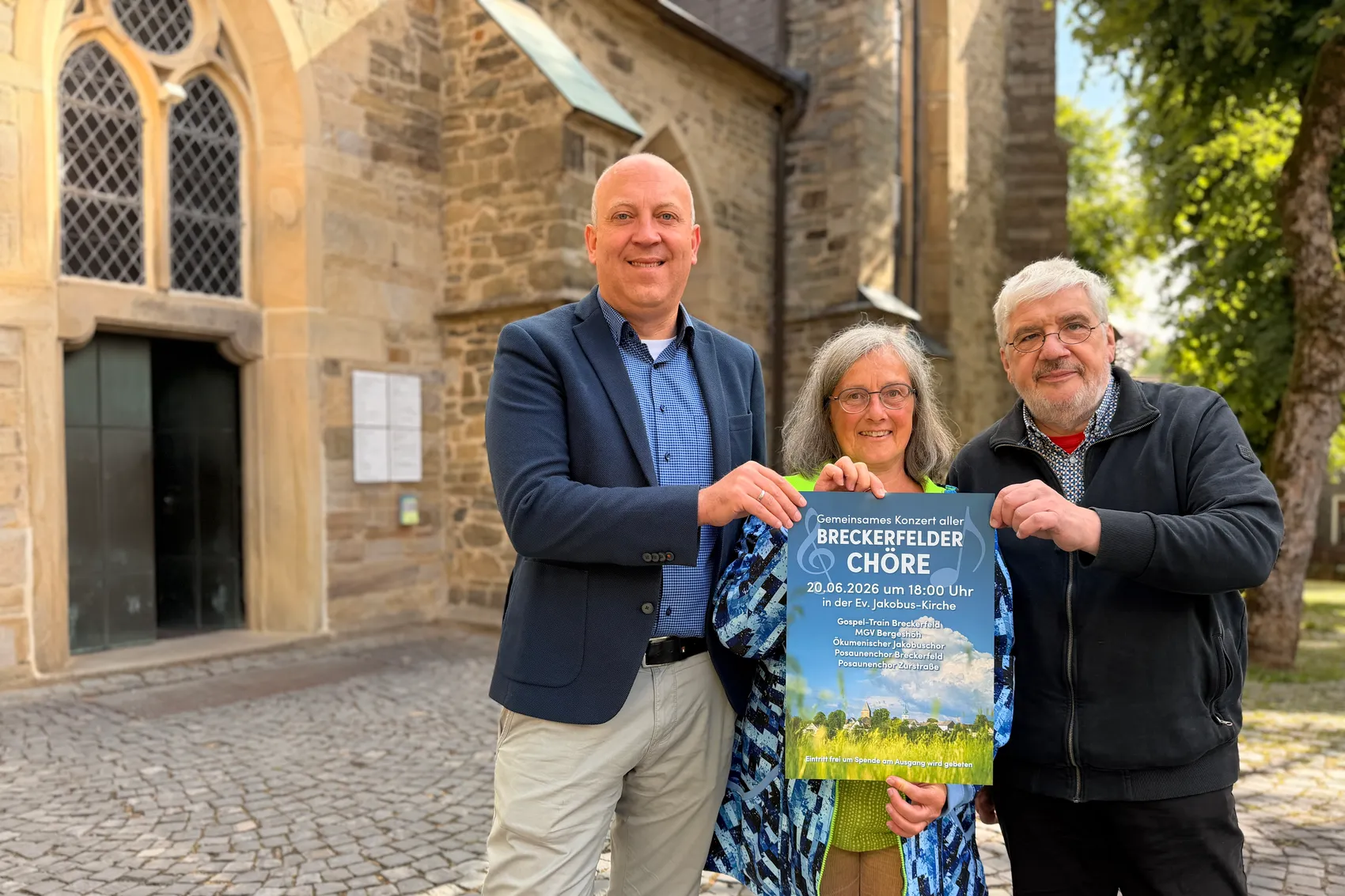 André Dahlhaus, Ute Slotkowski und Friedbert Schulz stehen vor der Jakobus-Kirche und halten eine Ankündigung für ein gemeinsames Konzert der Breckerfelder Chöre am 20. Juni um 18:00 Uhr.