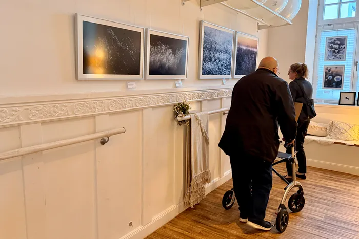 Ein Mann mit Gehhilfe und eine Frau betrachten Kunstwerke an einer weißen Wand in einem hellen Raum. Drei gerahmte Schwarz-Weiß-Fotografien hängen nebeneinander über einer verzierten Wandleiste.