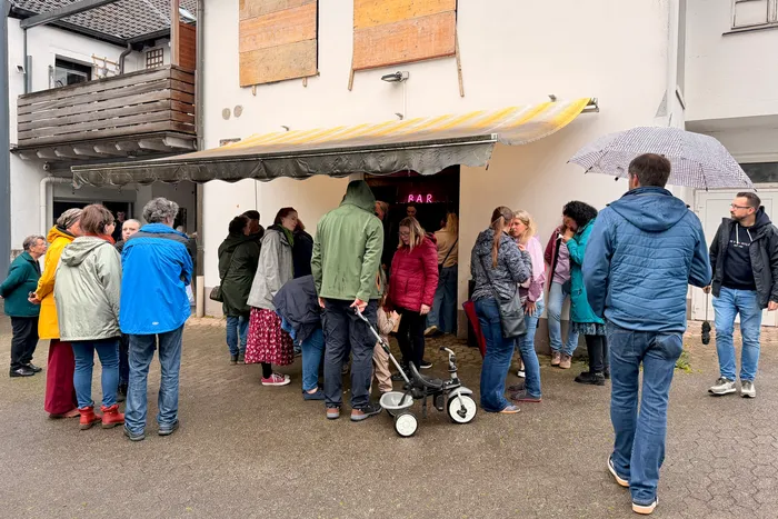 Menschen stehen unter einem Vordach und warten im Regen auf Einlass zu einer Veranstaltung. Über dem Eingang ist ein Leuchtschild mit der Aufschrift 