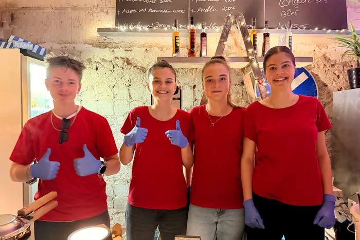 Vier junge Frauen in roten T-Shirts und blauen Handschuhen stehen lächelnd hinter einem Tresen. Im Hintergrund sind Getränke und eine Tafel mit Preisangaben zu sehen.