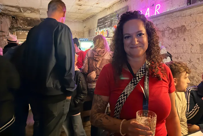A woman with curly dark hair smiles at the camera while holding a glass in a crowded bar. People are visible in the background, some standing at the bar, others walking through the room.