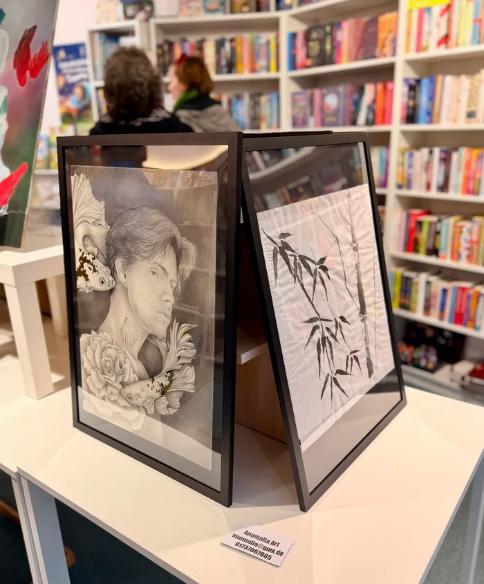 Zwei gerahmte Schwarz-Weiß-Zeichnungen stehen auf einem Tisch vor einem Bücherregal. Links ist ein Porträt einer Person mit Blumen, rechts eine Darstellung von Vögeln auf Zweigen.