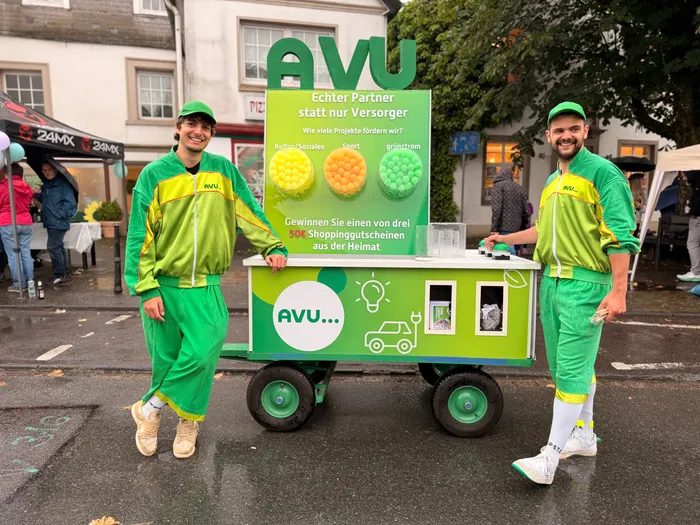 Zwei junge Männer in grünen AVU-Jacken und -Mützen stehen lächelnd neben einem mit Werbung versehenen Rollwagen. Der Wagen zeigt bunte Kugeln und bewirbt Shoppinggutscheine.