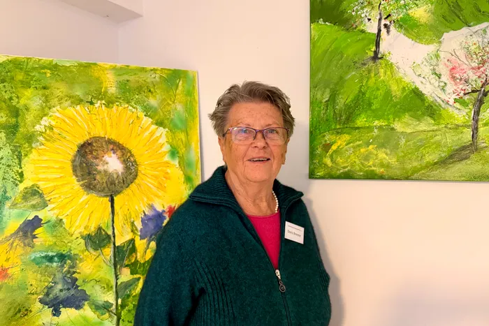 Eine Frau mit kurzem, grauem Haar und Brille lächelt in die Kamera. Im Hintergrund hängen zwei farbenfrohe Gemälde, eines mit einer großen Sonnenblume und das andere mit blühenden Obstbäumen.