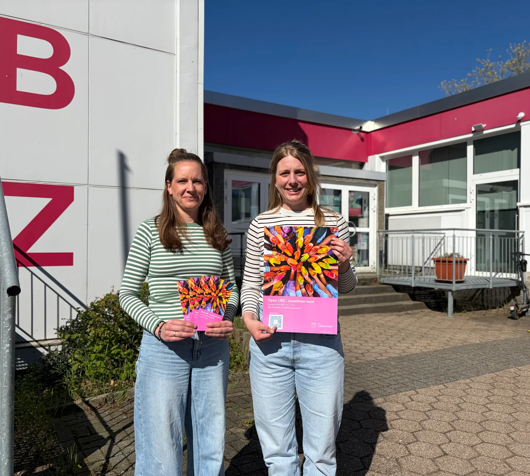 Zwei Frauen halten farbenfrohe Programmhefte mit dem Titel „Open Litz – zusammen bunt“ vor einem Gebäude mit einem großen roten „B“ und „Z“.