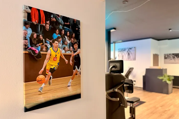 Ein gerahmtes Foto an einer weißen Wand zeigt zwei Basketballspieler in Aktion vor einem unscharfen Publikum. Im Vordergrund des Bildes befindet sich ein Bürobereich mit einem Schreibtisch und einem Stuhl.